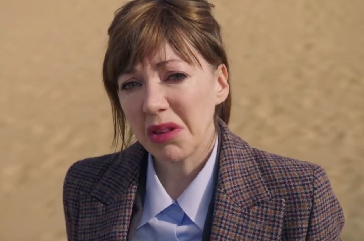 Philomena Cunk in "Cunk on Earth" outdoors wearing a plaid blazer and light blue shirt, looking directly at the camera with a disgusted expression