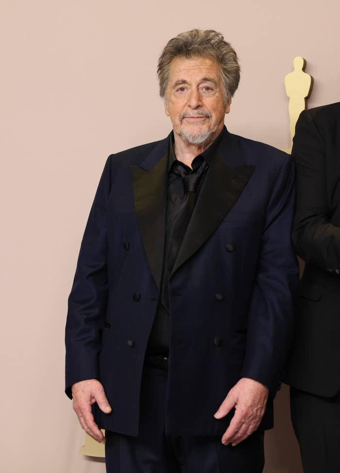 A person in a dark suit with a double-breasted jacket stands on a formal event backdrop featuring award statue silhouettes