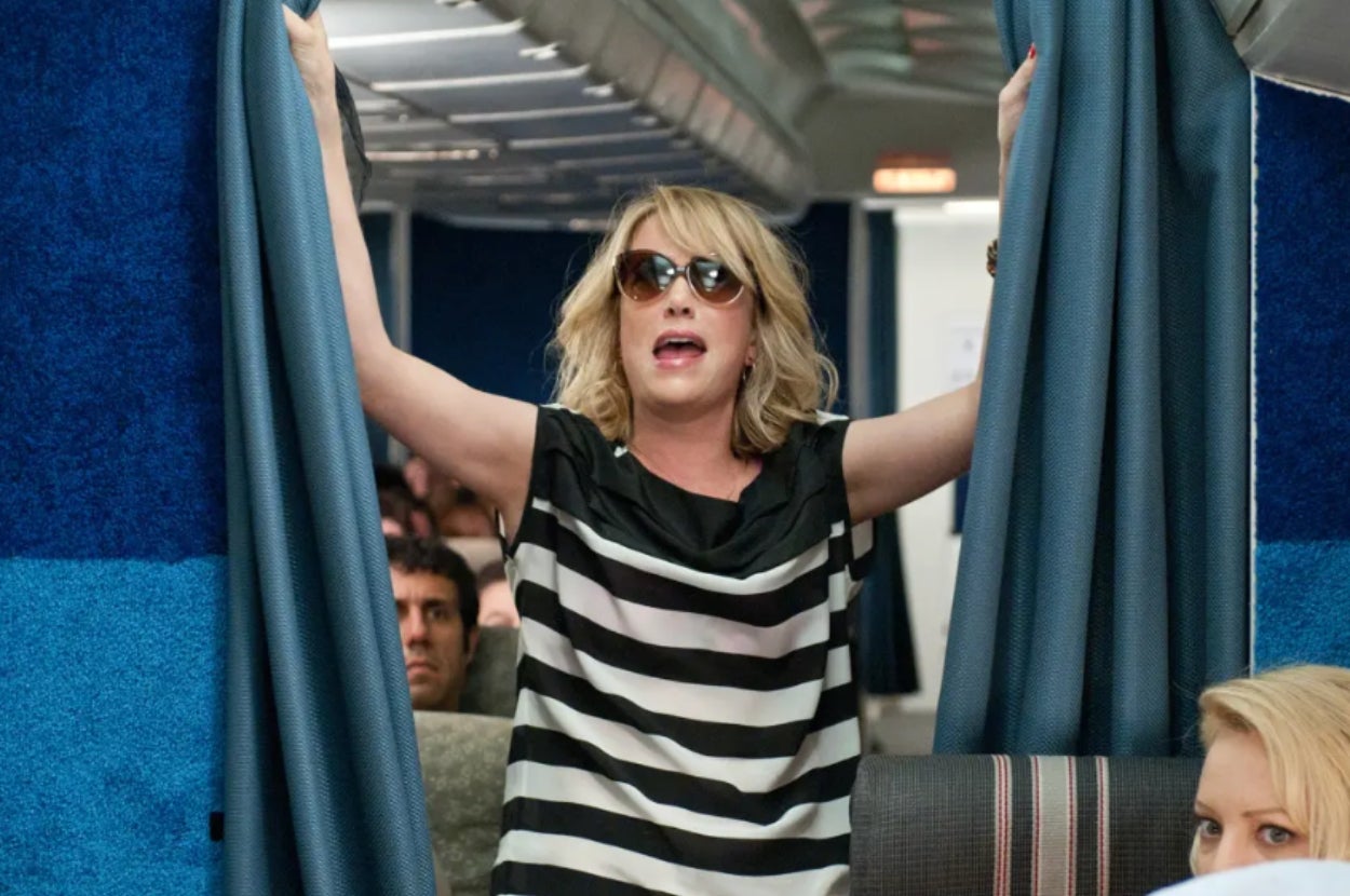 A person on a plane dramatically holds open curtains, wearing sunglasses and a black-and-white striped shirt. Passengers are visible in the background