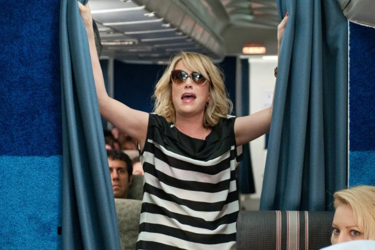 A person on a plane dramatically holds open curtains, wearing sunglasses and a black-and-white striped shirt. Passengers are visible in the background
