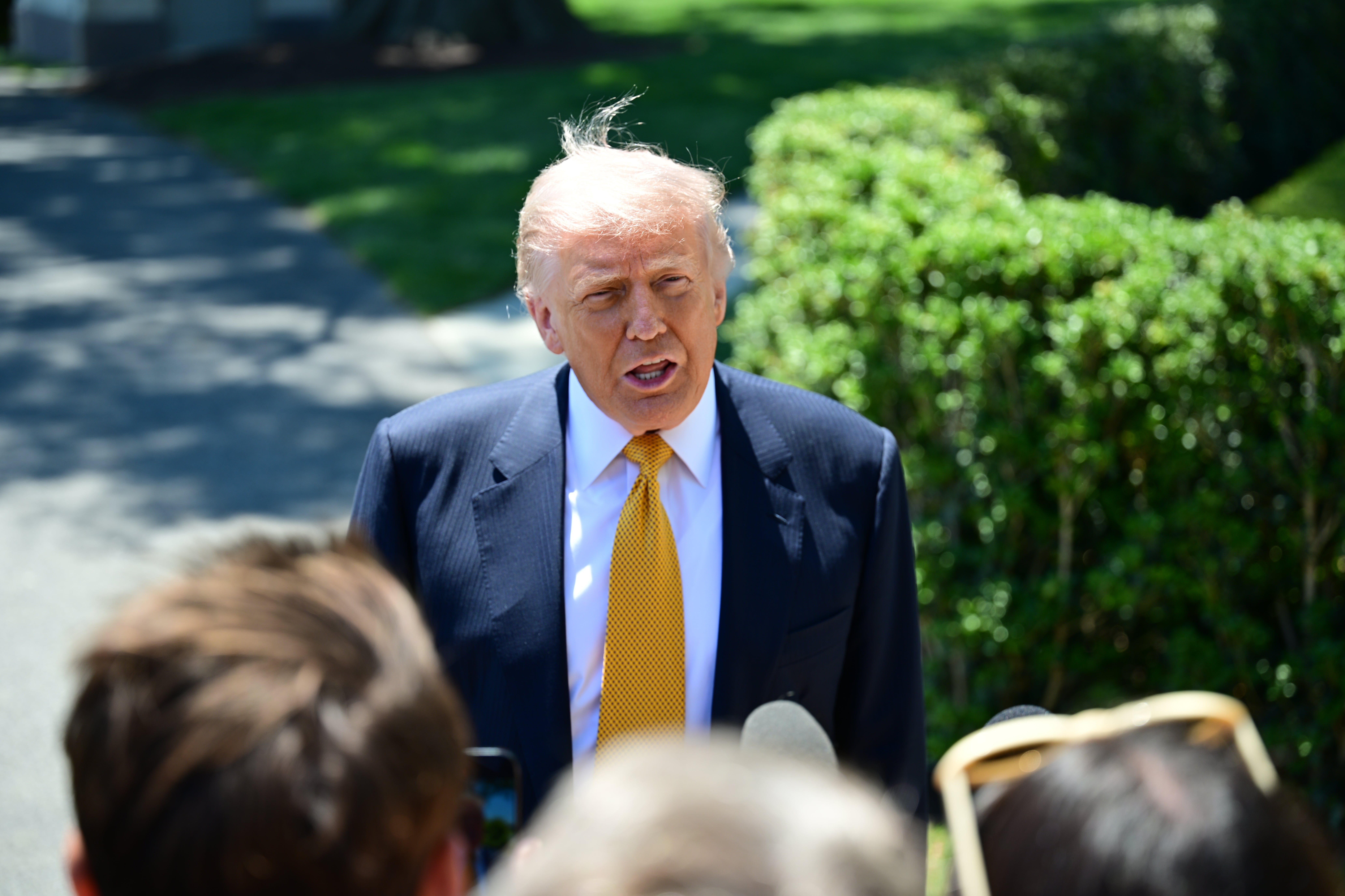 A person in a suit and tie speaking to reporters outdoors in front of a hedge. Microphones are visible in the foreground