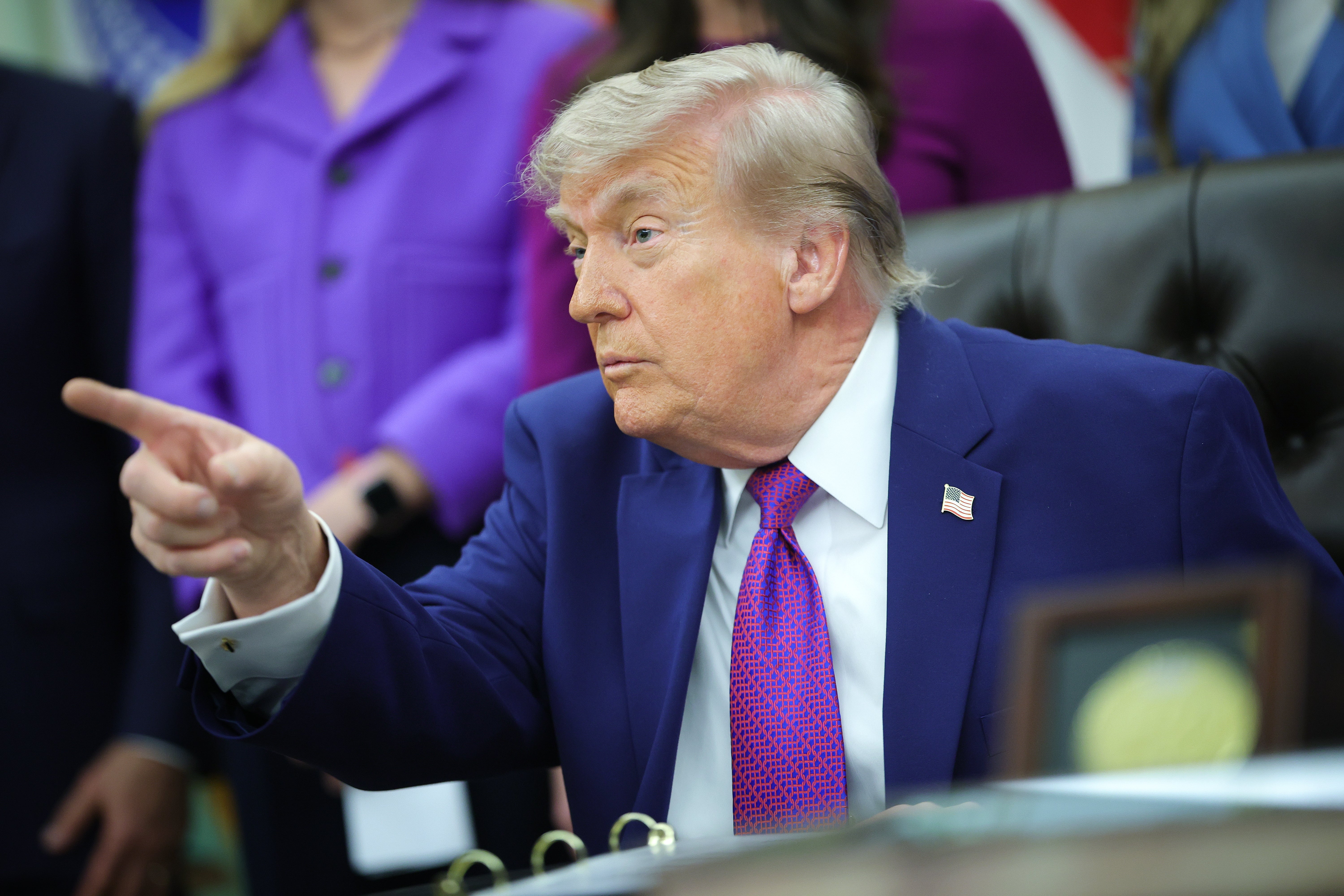 Public figure gesturing while seated in an office setting, wearing a suit with a tie. People are visible in the background