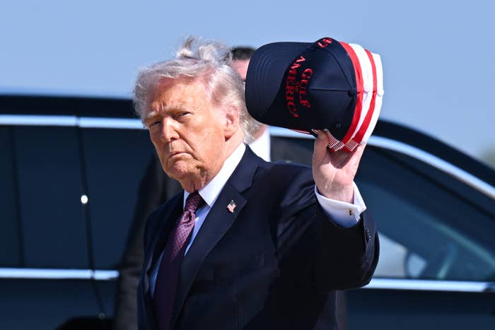 Person in suit holding a "Save America" hat, beside a vehicle. Hair is windblown. Location and event are unspecified