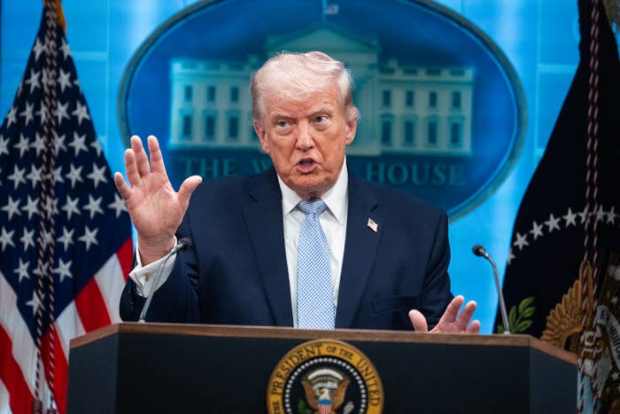 A person in a suit stands at a lectern with a presidential seal, gesturing while speaking, with flags and a blurred backdrop of a government building