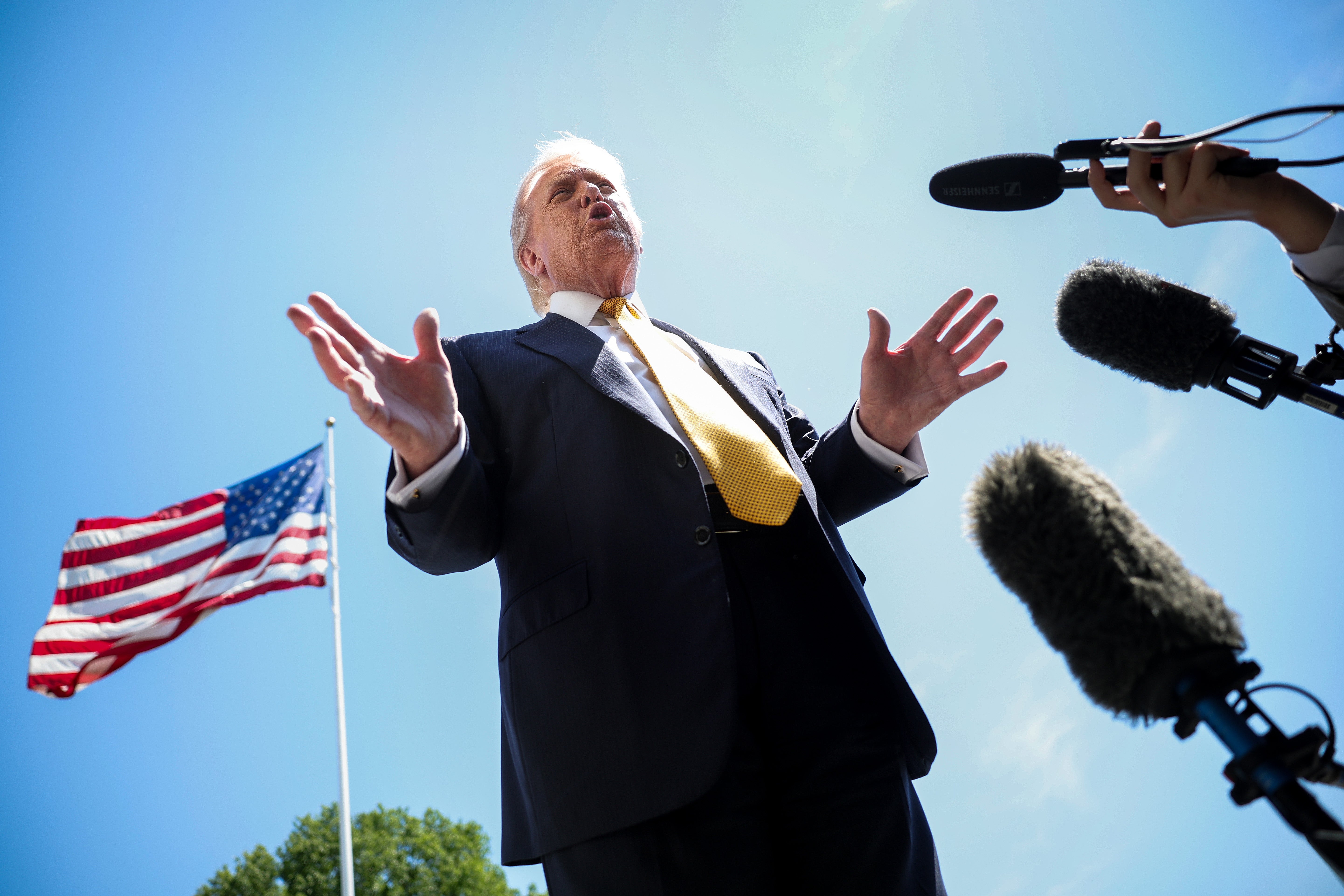 Person in a suit speaks to reporters with microphones outside, near an American flag, likely making a public statement or answering questions