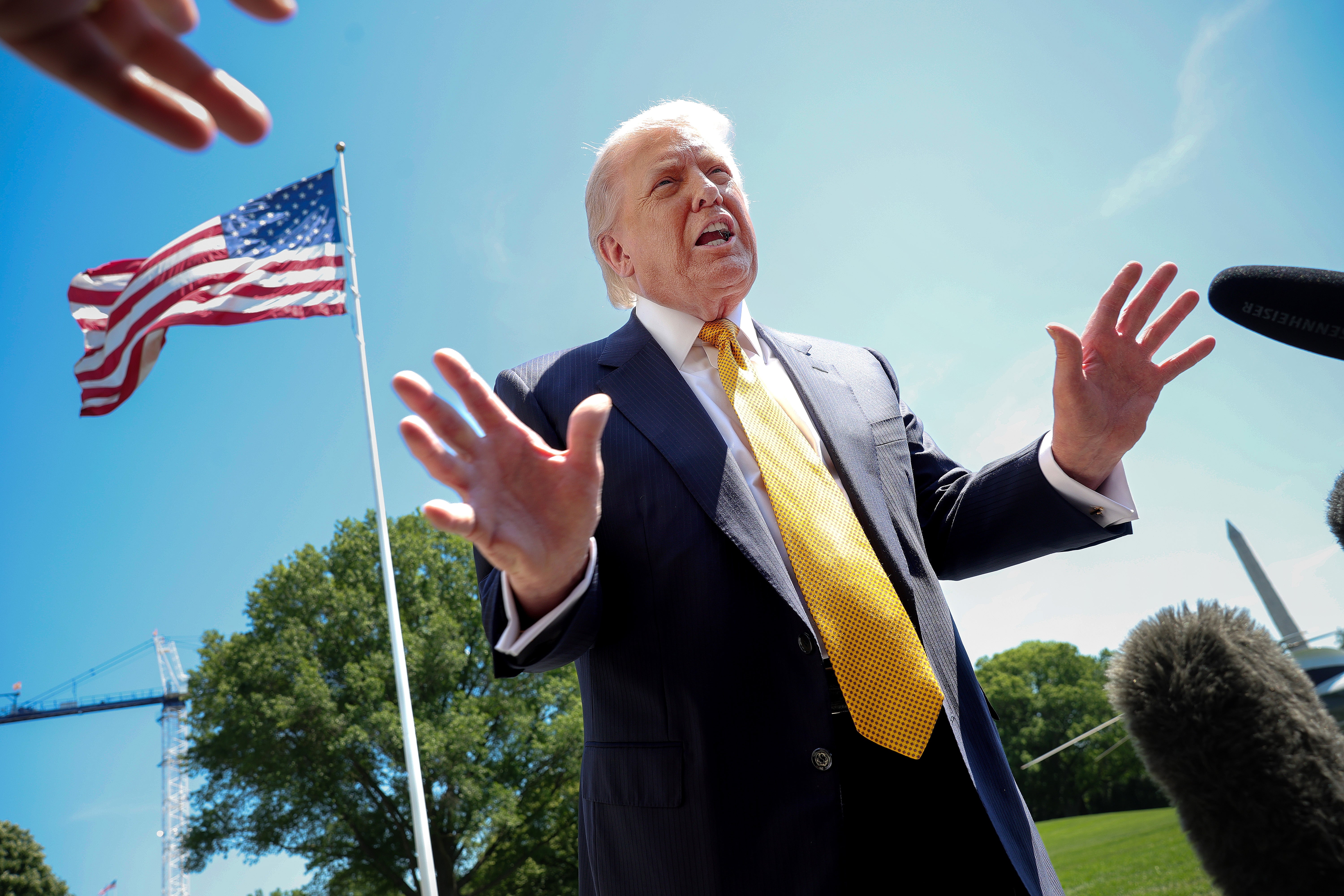 A man in a suit and yellow tie speaks passionately outdoors near a U.S. flag, surrounded by microphones