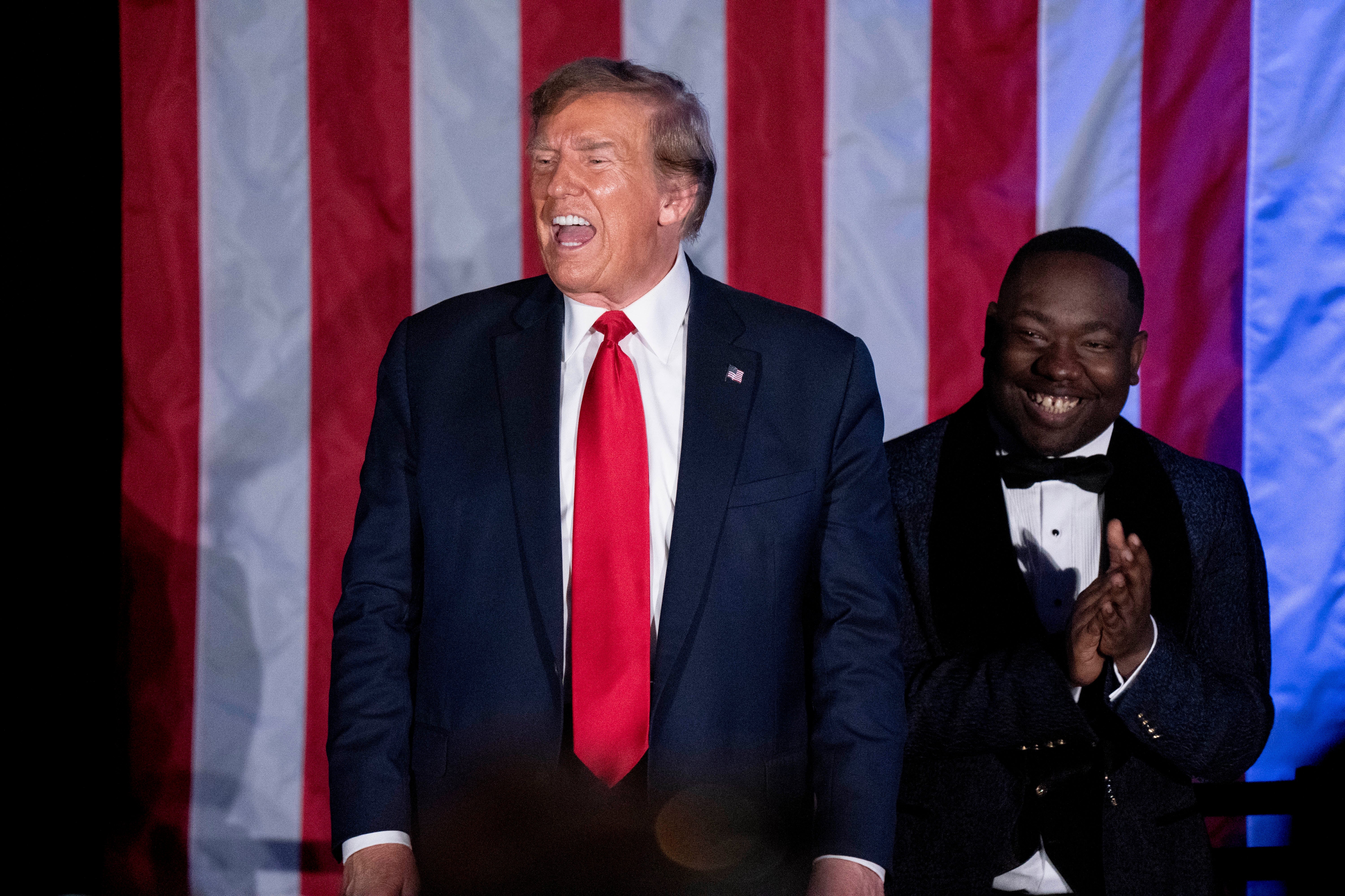 Two people in formal attire stand in front of a U.S. flag backdrop, one smiling broadly and clapping