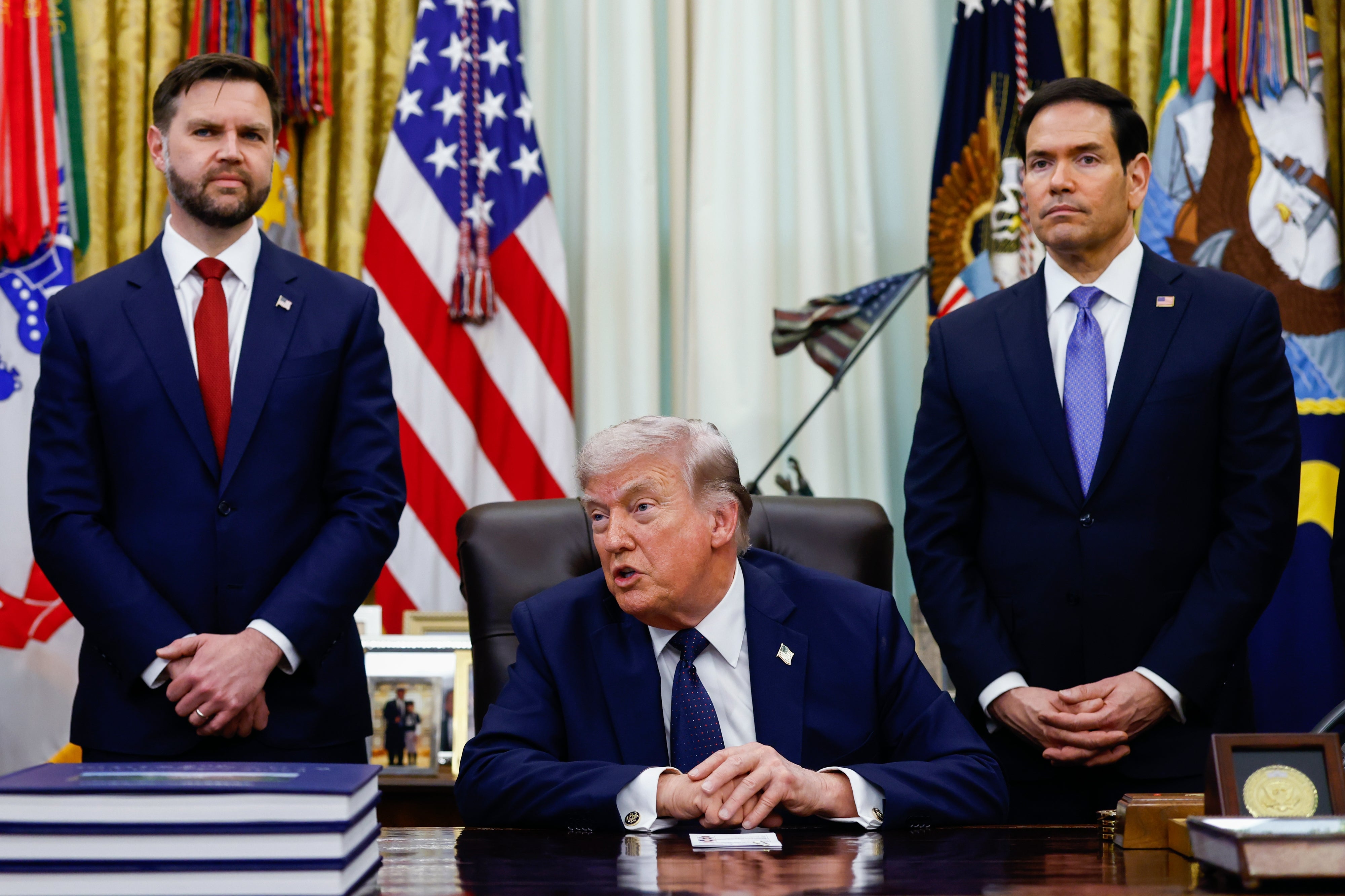 Three men in suits stand and sit around a desk with flags in the background, suggesting a formal or official event