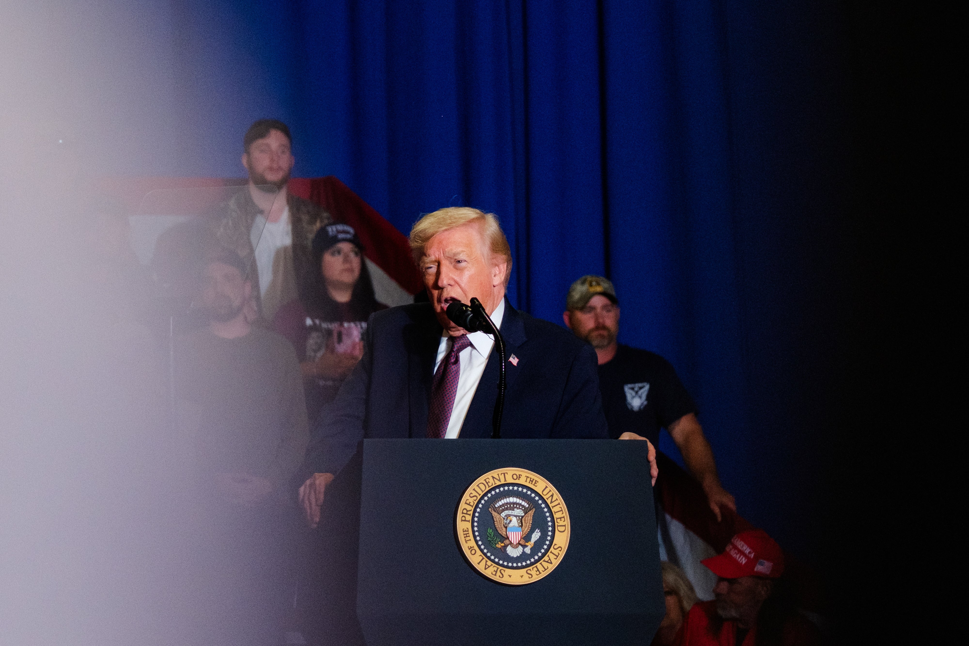 Person speaking at a podium with a presidential seal, in front of a crowd and blue backdrop
