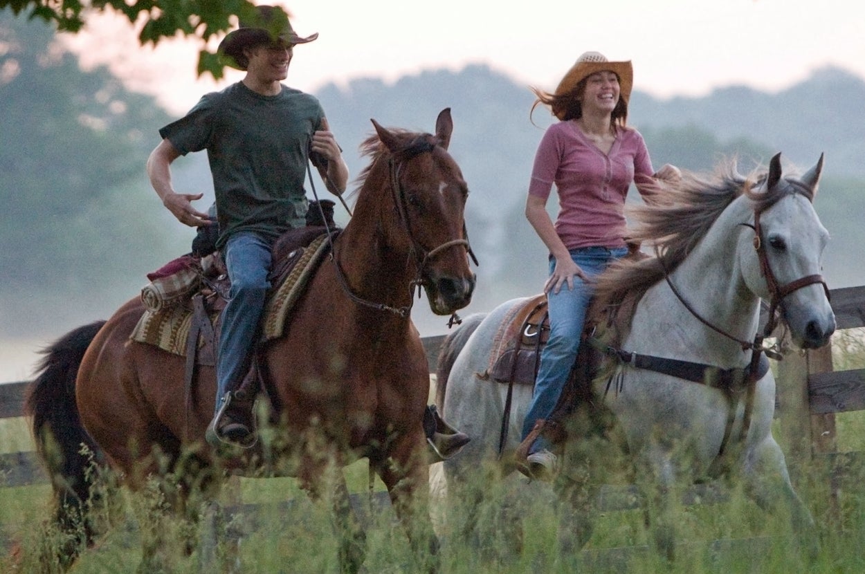 Two people wearing hats ride horses on a grassy path, enjoying a leisurely outdoor activity