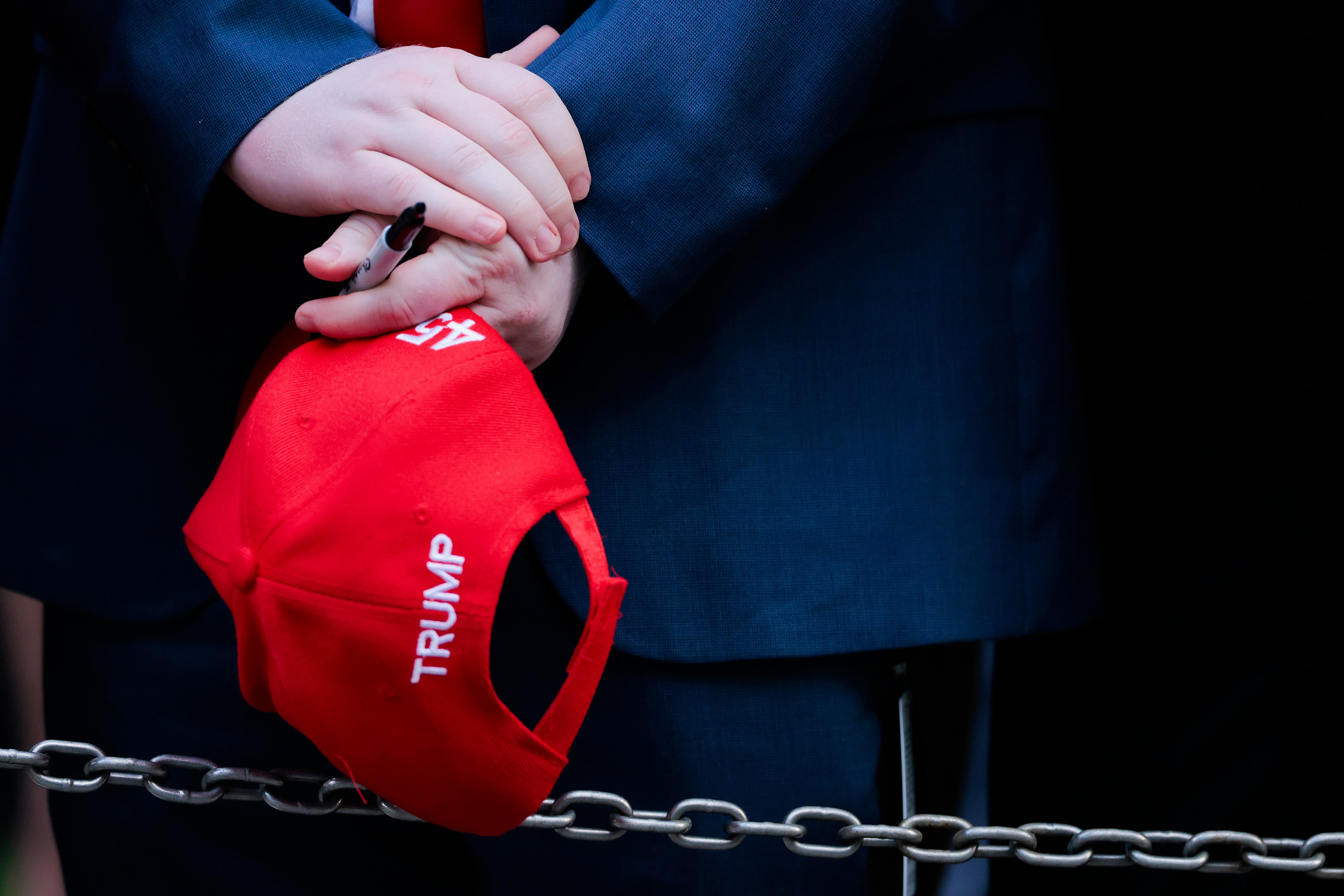 Person holding a red "Trump" cap, wearing a dark suit, behind a chain barrier. Hands crossed with a pen visible