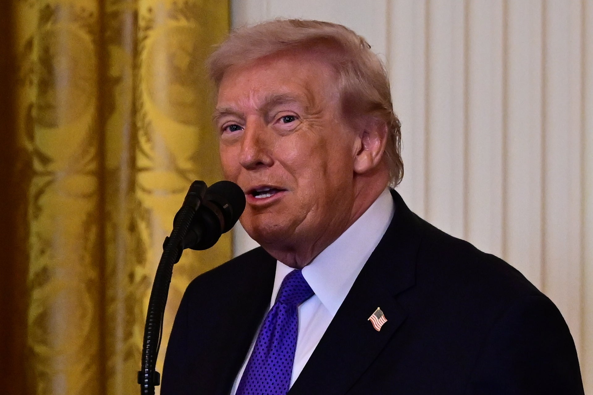 A person speaking into a microphone at a formal event, wearing a suit and tie, with a flag pin on their lapel