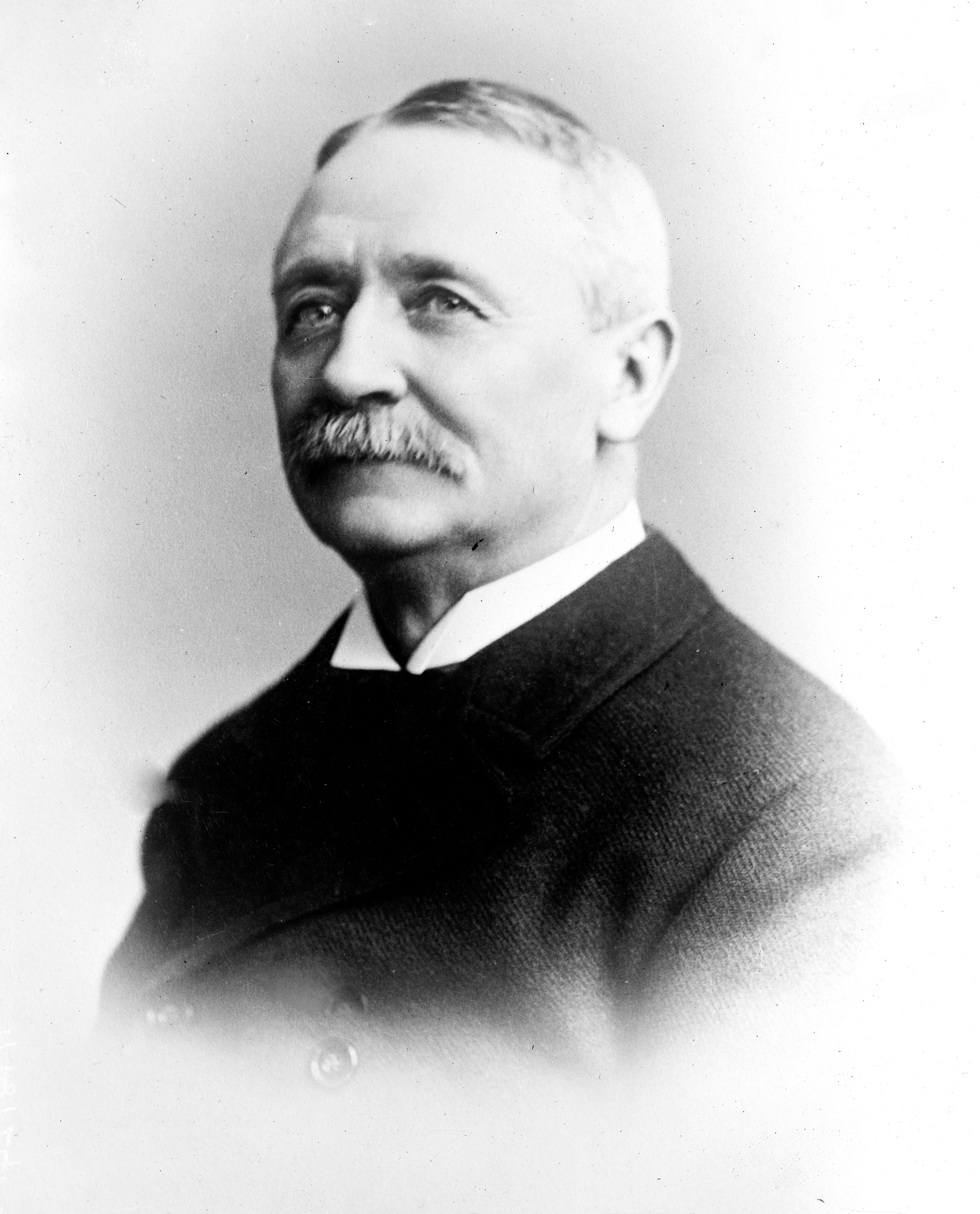 A distinguished older gentleman with a mustache, dressed in a formal dark suit, poses in a classic portrait style