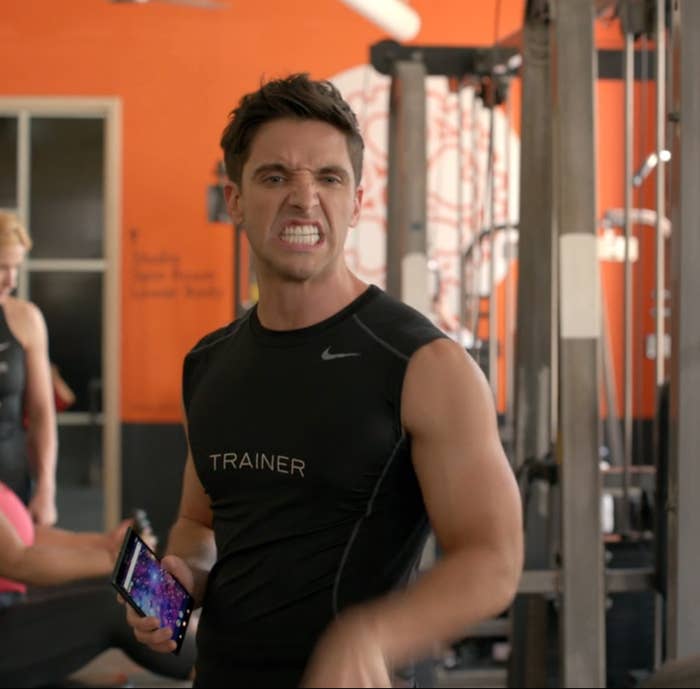 Man successful  gym holding phone, wearing achromatic  sleeveless garment  labeled "Trainer," making aggravated  expression. Gym instrumentality  disposable   successful  background
