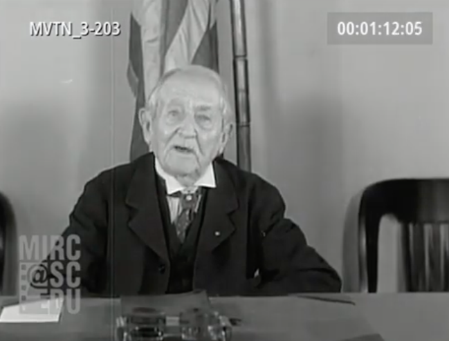 An elderly man sits at a table, speaking, with a flag visible in the background. 