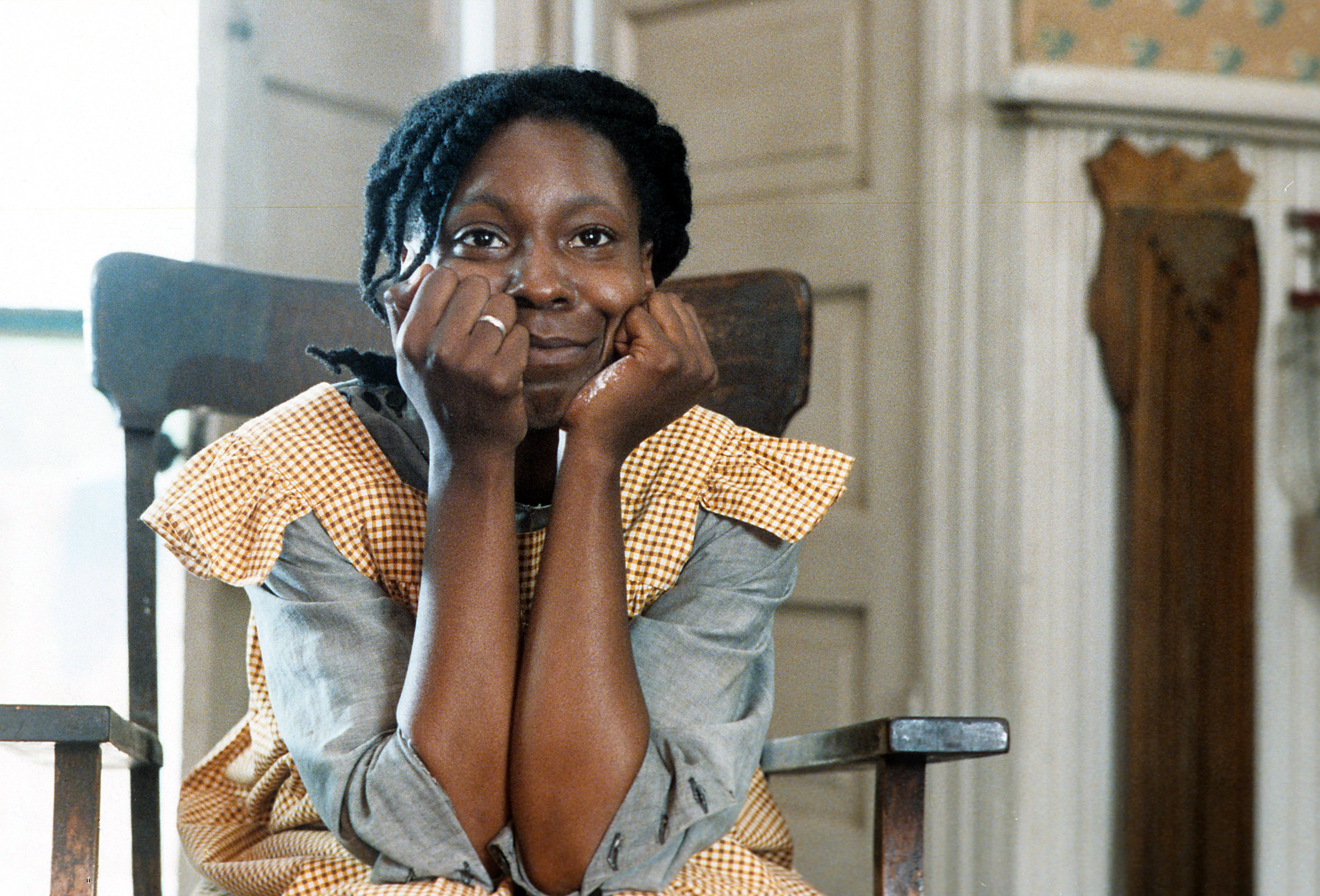 Actor sits in a vintage chair, wearing a gingham dress, with their chin resting on their hands, looking contemplative in a classic film setting