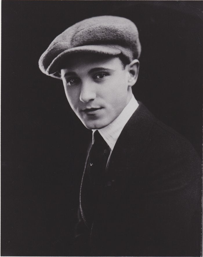 A man in early 20th-century attire with a flat cap and suit poses for a portrait