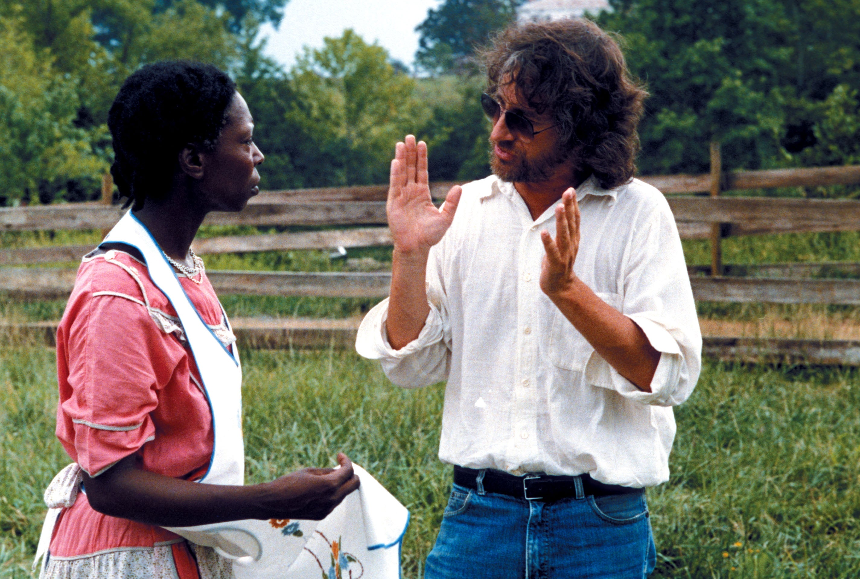 A person in period costume talks with another in casual modern clothing outdoors, gesturing animatedly