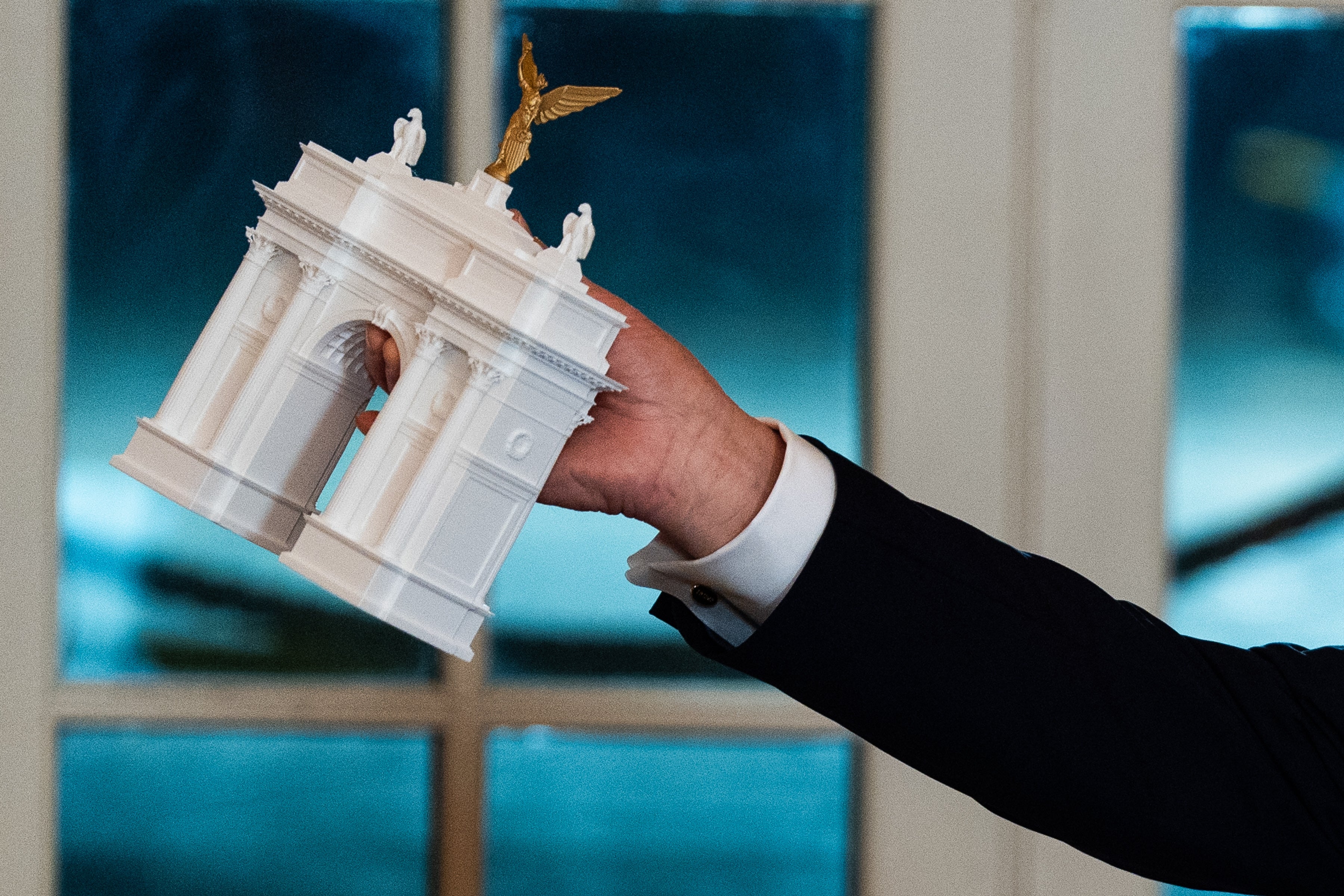 Person holding a elaborate miniature replica of a classical-style monument with columns and a winged fig connected top
