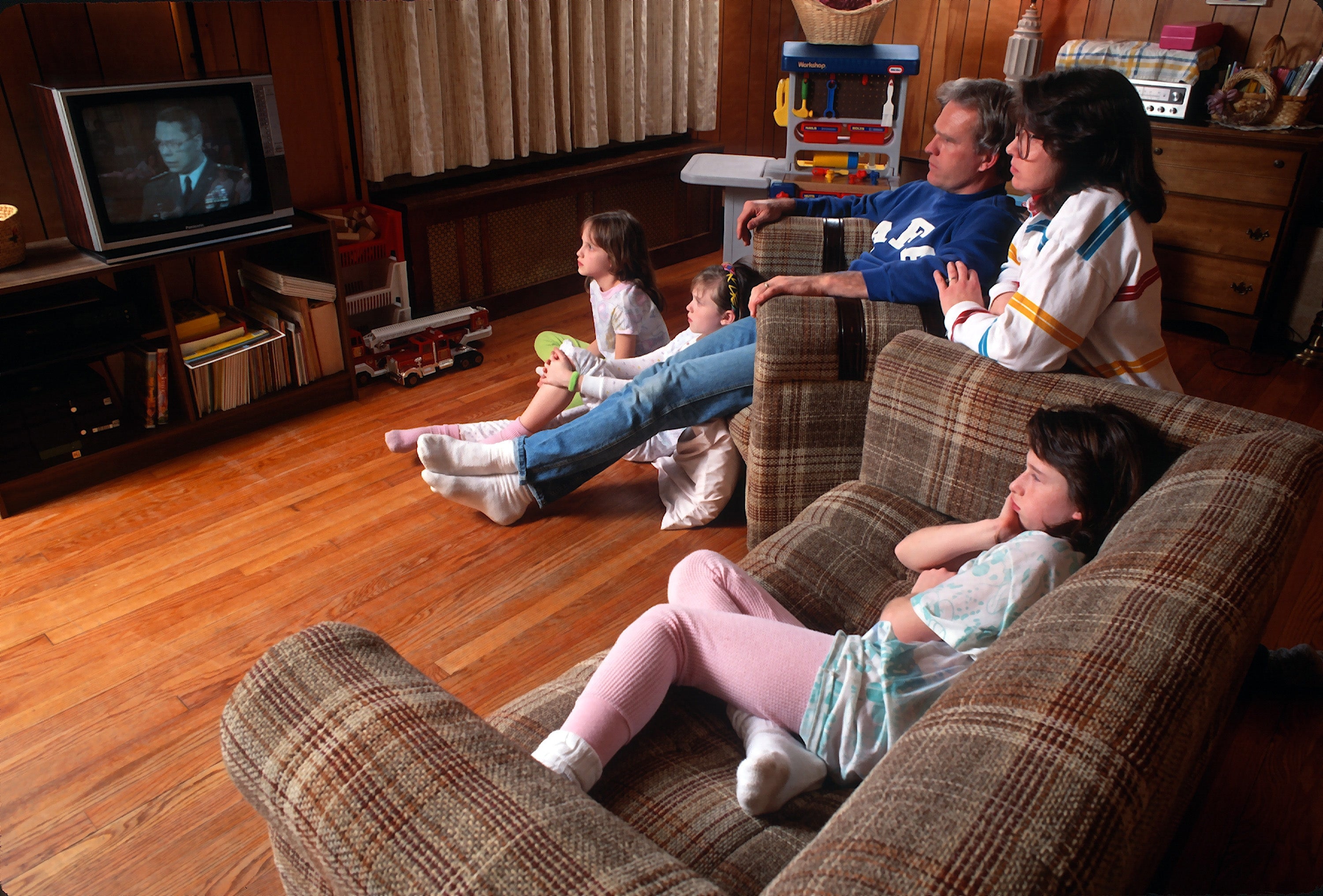 Family of five sits on couches, watching TV together in a cozy living room. Children seem engaged with the program