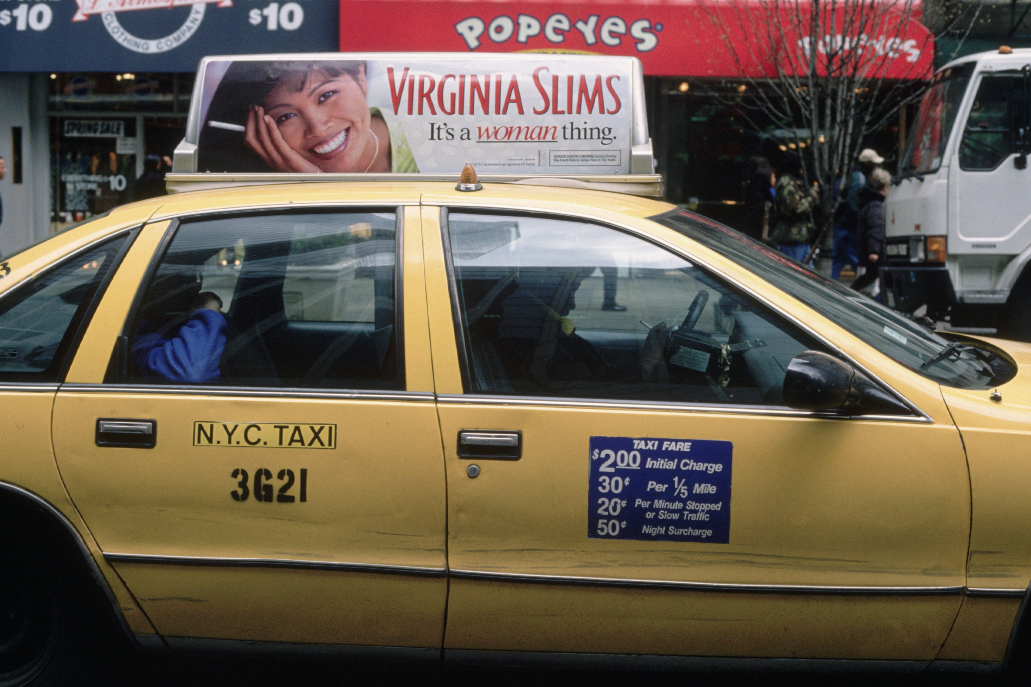 Yellow NYC taxi with a Virginia Slims ad on top. Passenger's hand visible through the window. Pricing details are displayed on the door