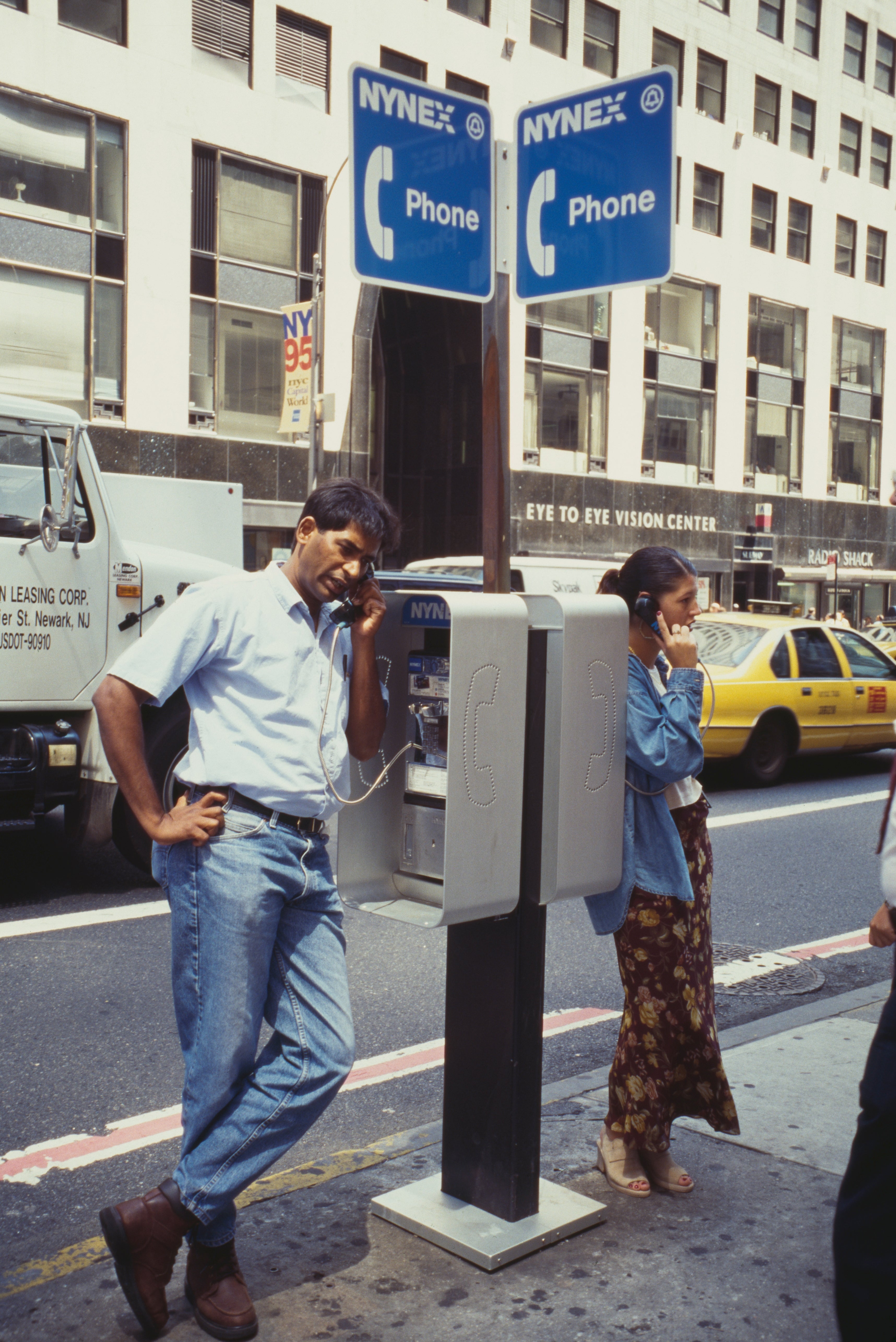 Two people use payphones on a busy city street. One is leaning casually, and the other is standing near a taxi