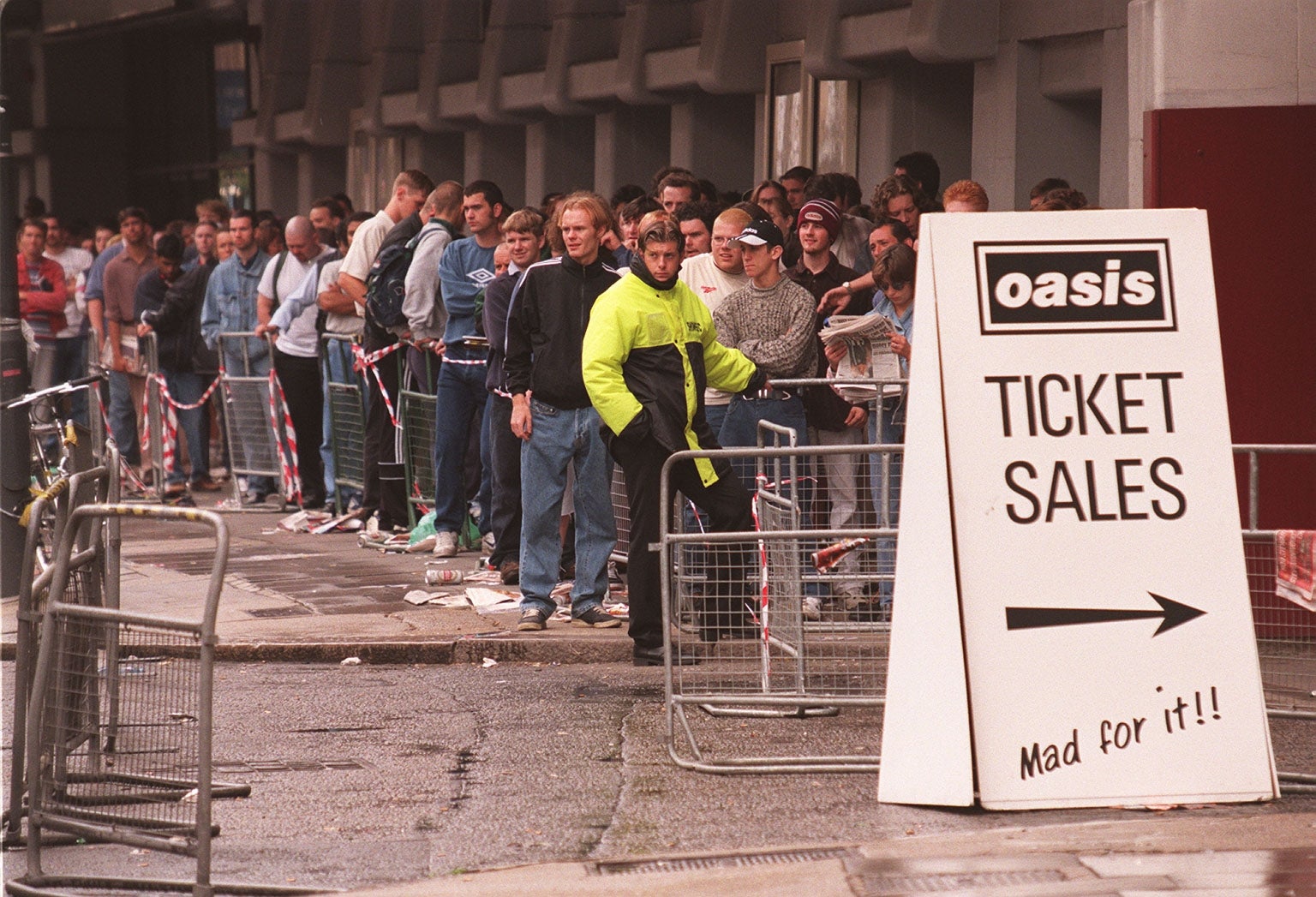People line up outside a building for Oasis concert tickets; a sign reads "Ticket sales. Mad for it!"