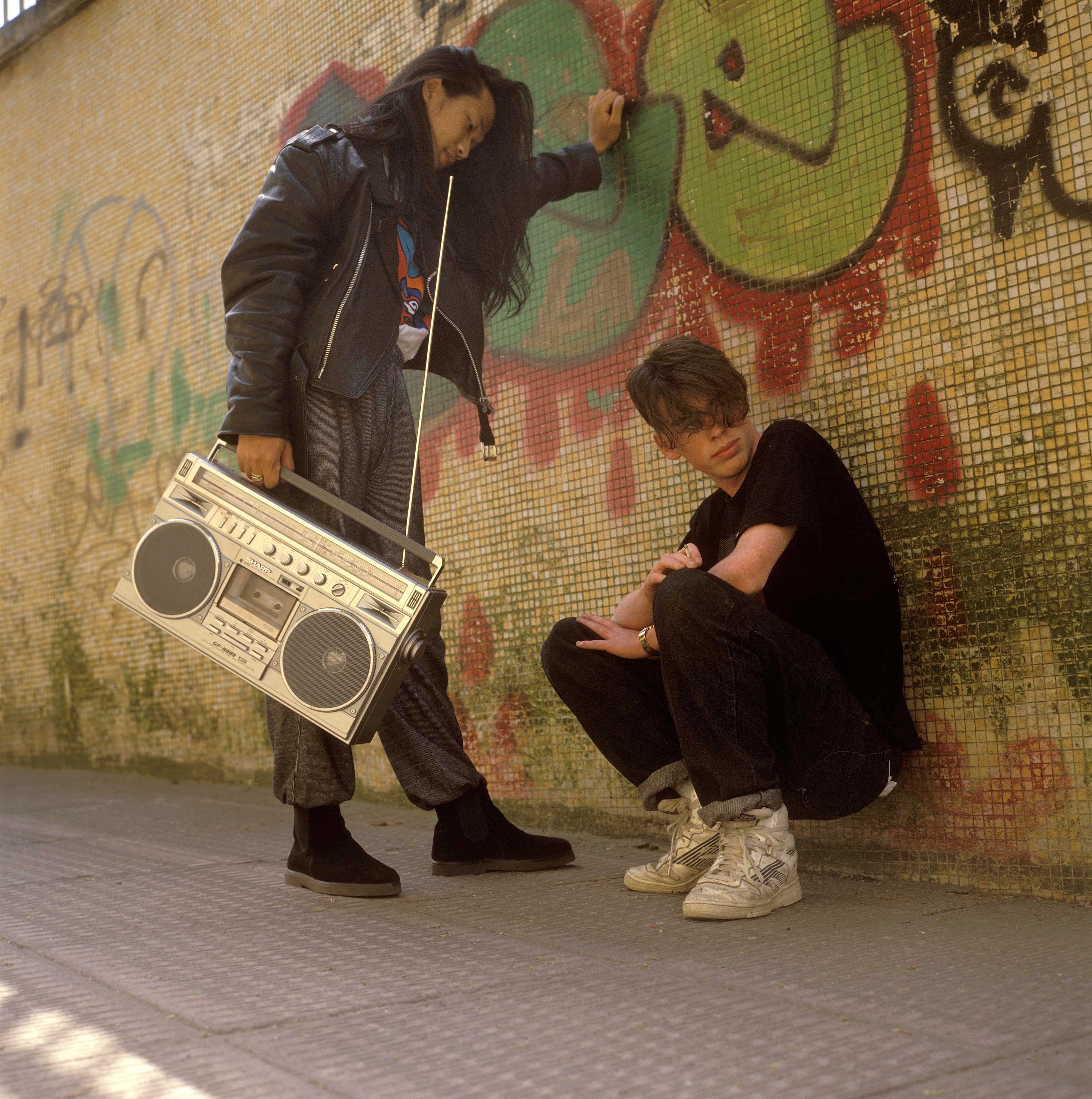 Two people in casual 1990s streetwear pose with a large boombox against a graffiti-covered wall