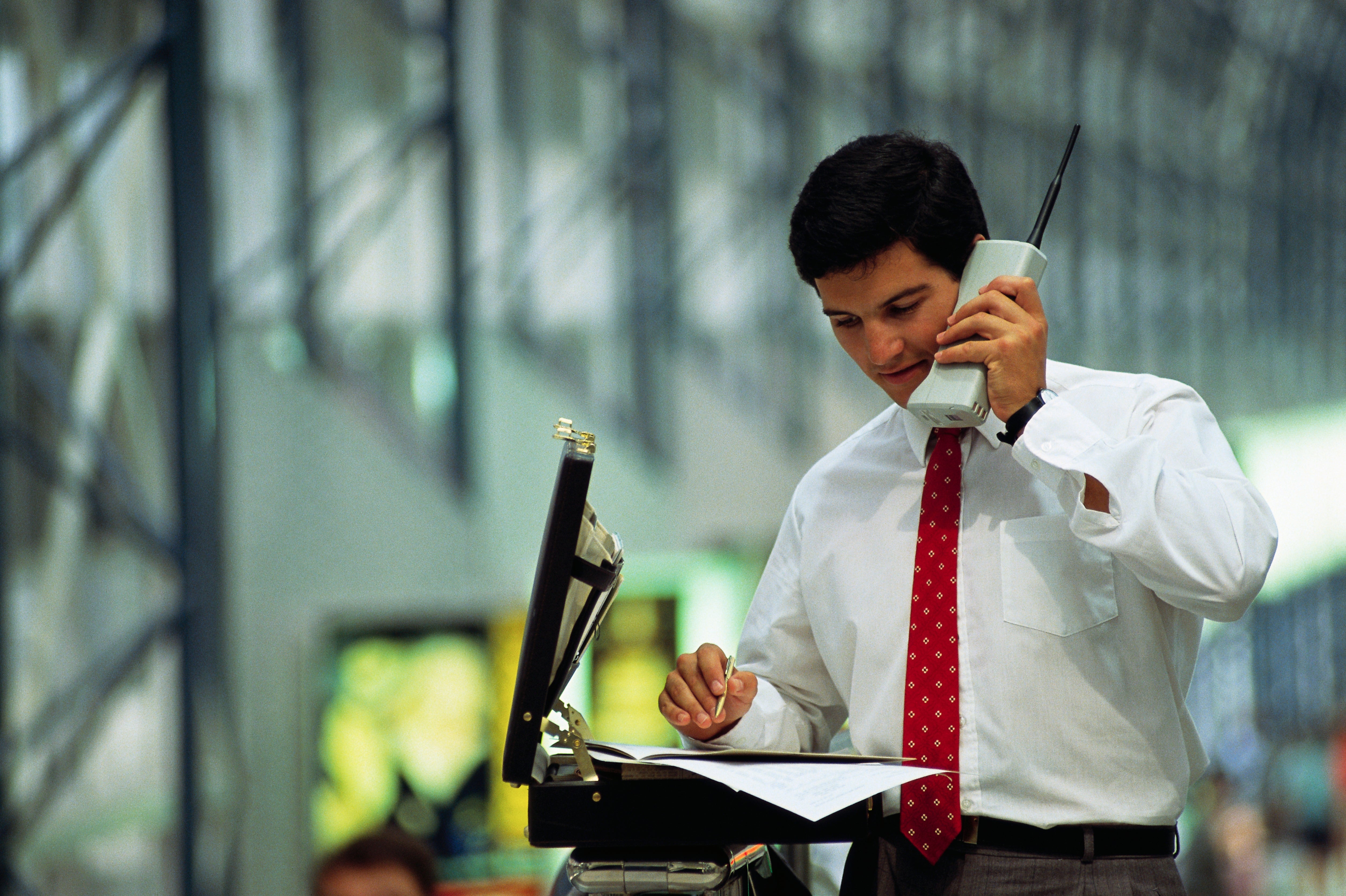 Person in a tie using a large cell phone, looking at papers on a briefcase in an office setting