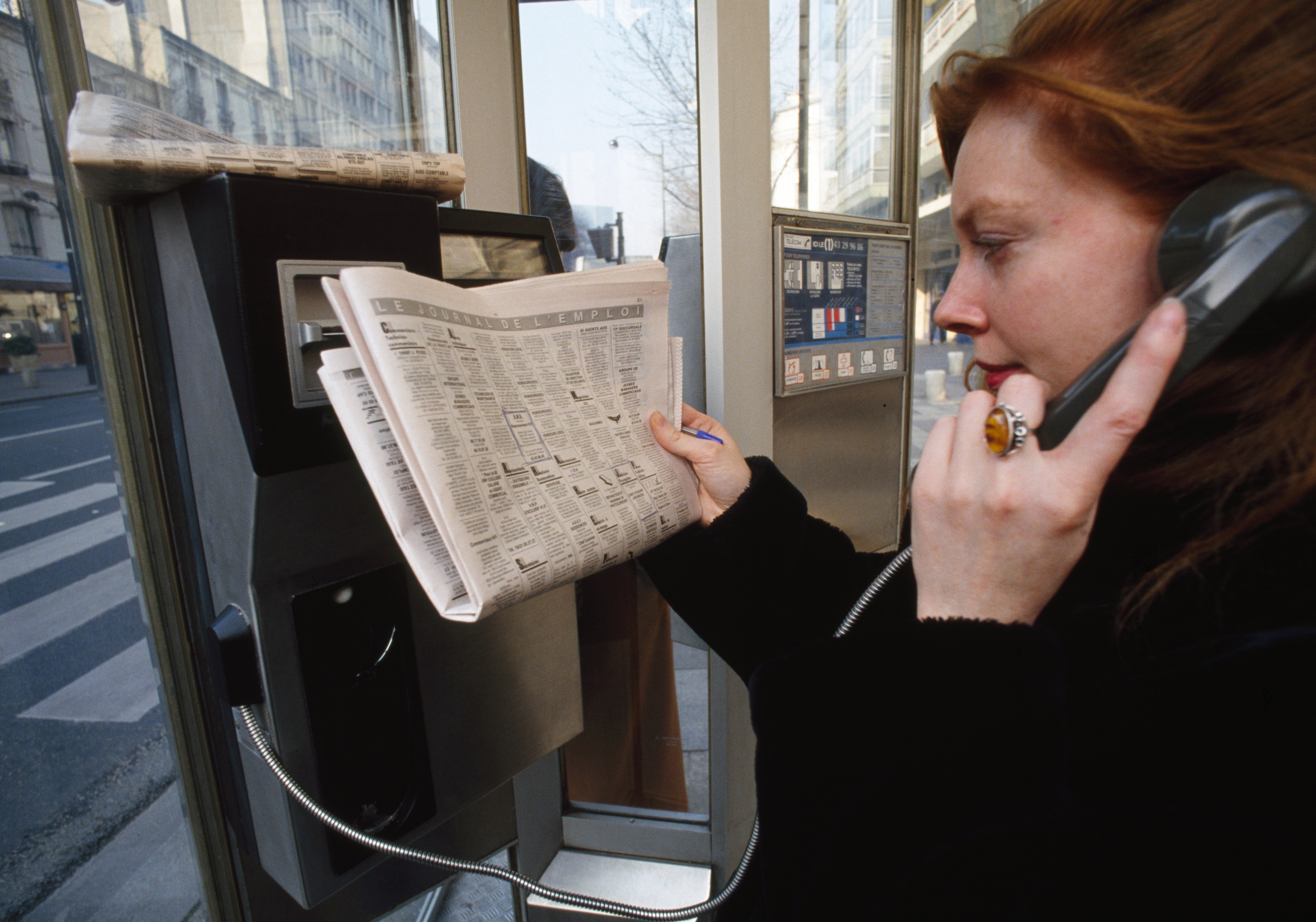 A person using a public phone booth while holding a newspaper, focused on a conversation
