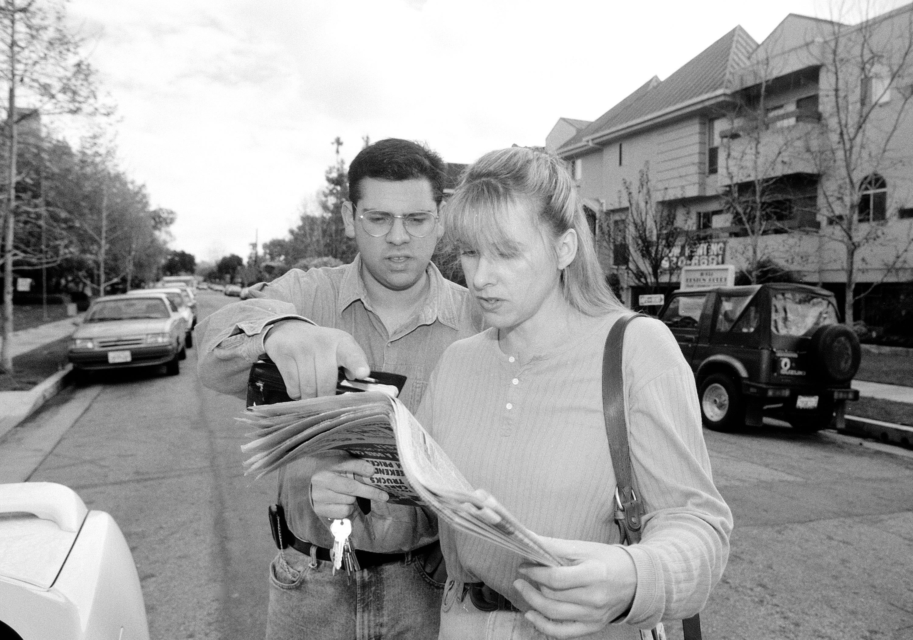 Two people stand on a street reading a newspaper. One holds a camera; they seem focused and engaged. Cars and houses line the background