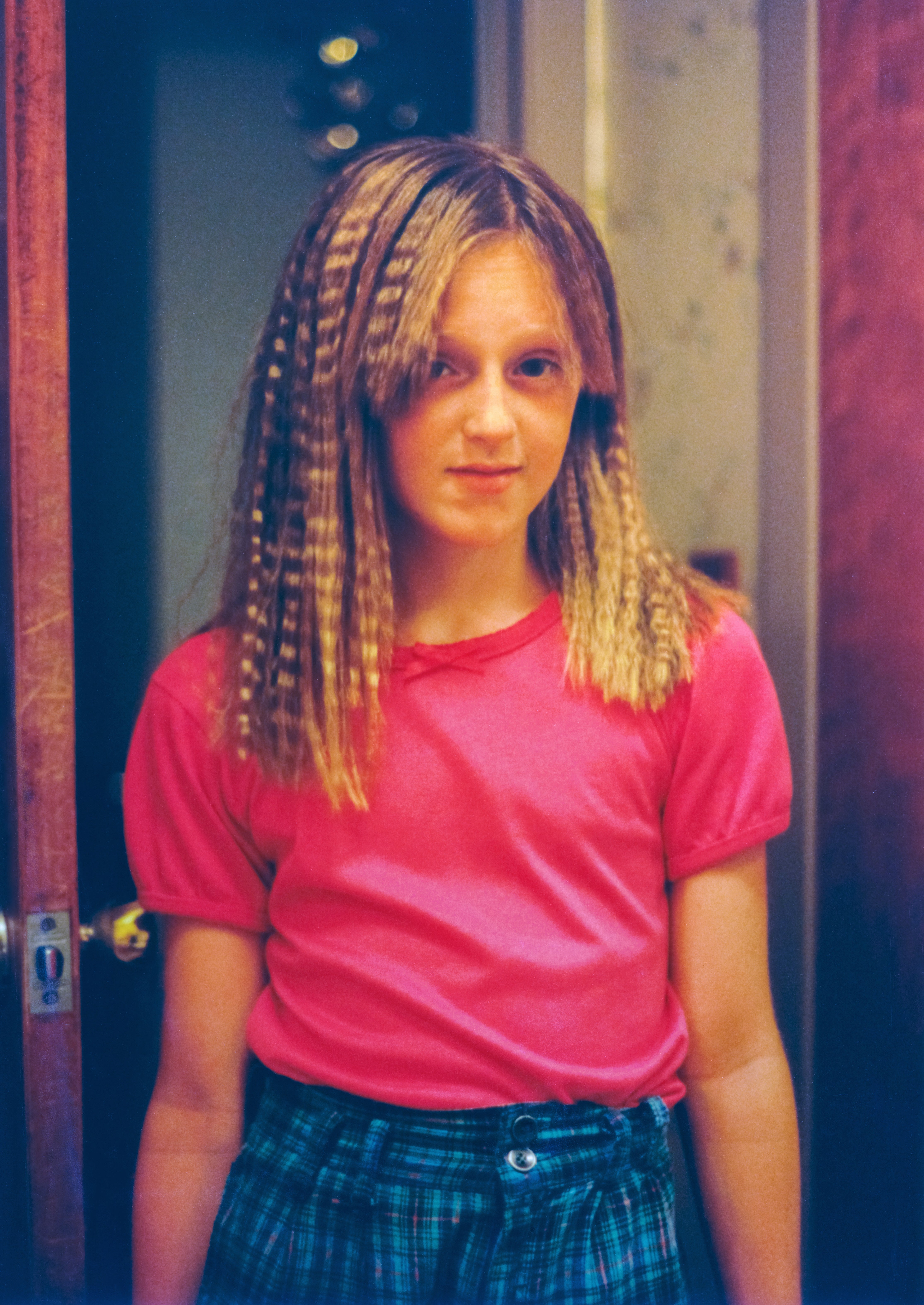A person with crimped hair wearing a pink top and patterned pants stands indoors, looking at the camera