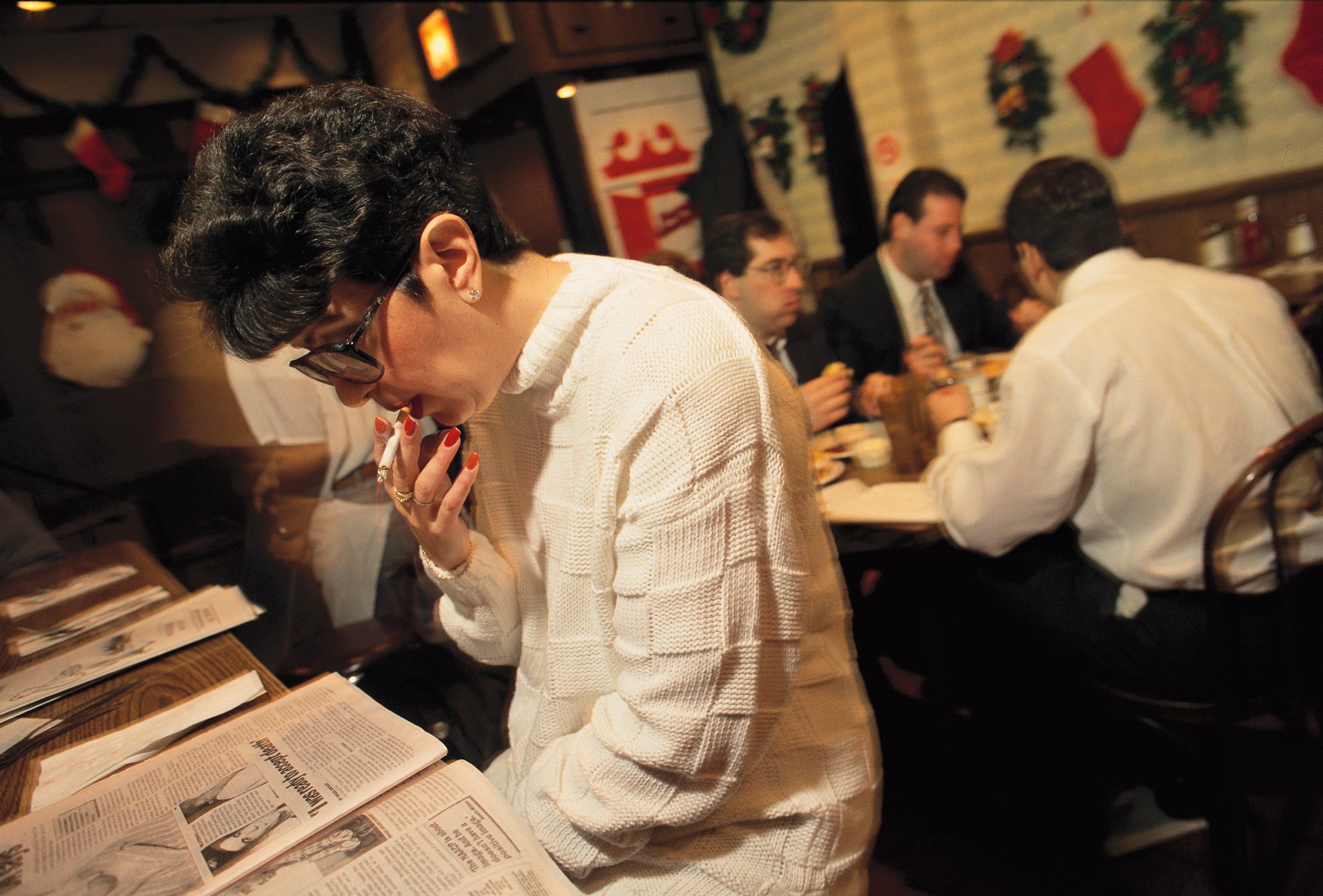 Person in a cozy restaurant reads a newspaper while smoking; people in the background are engaged in conversation at their table