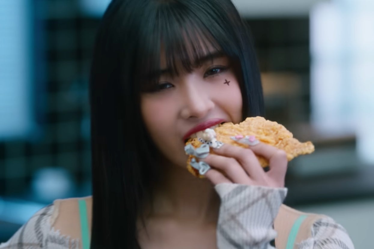 Sophia from "Katseye" in "Gnarly" music video biting crispy fried chicken, with star face sticker and adorned nails, smiling slightly