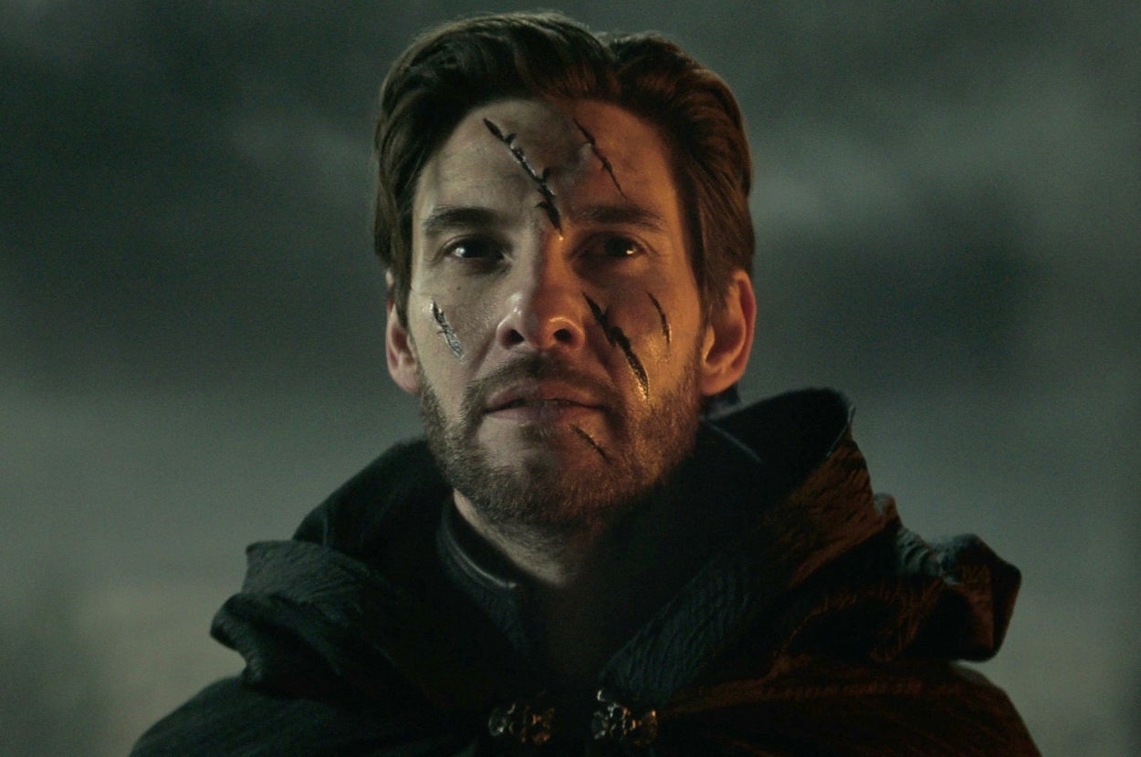 Benjamin Barnes as the Darkling in "Shadow and Bone" with a cloak and facial scars stares ahead in a dramatic, moody setting