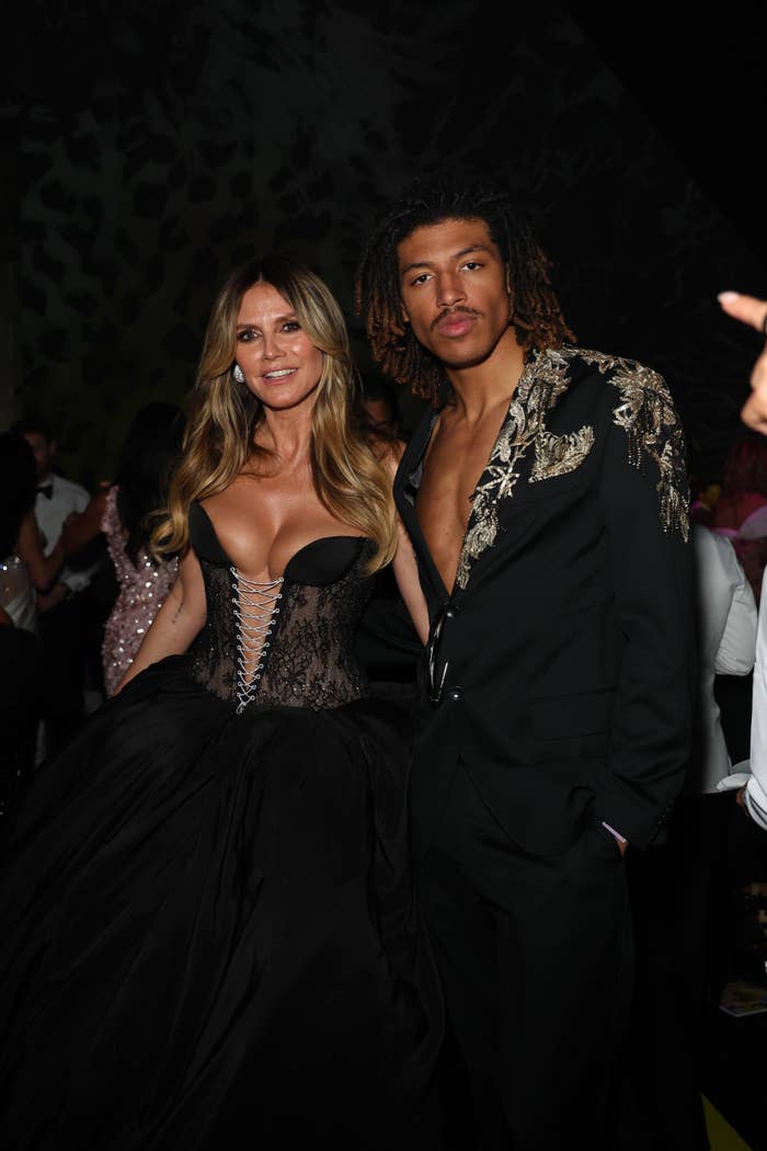 Two people pose together at an event; the woman in a strapless corset gown and the man in an embroidered suit jacket