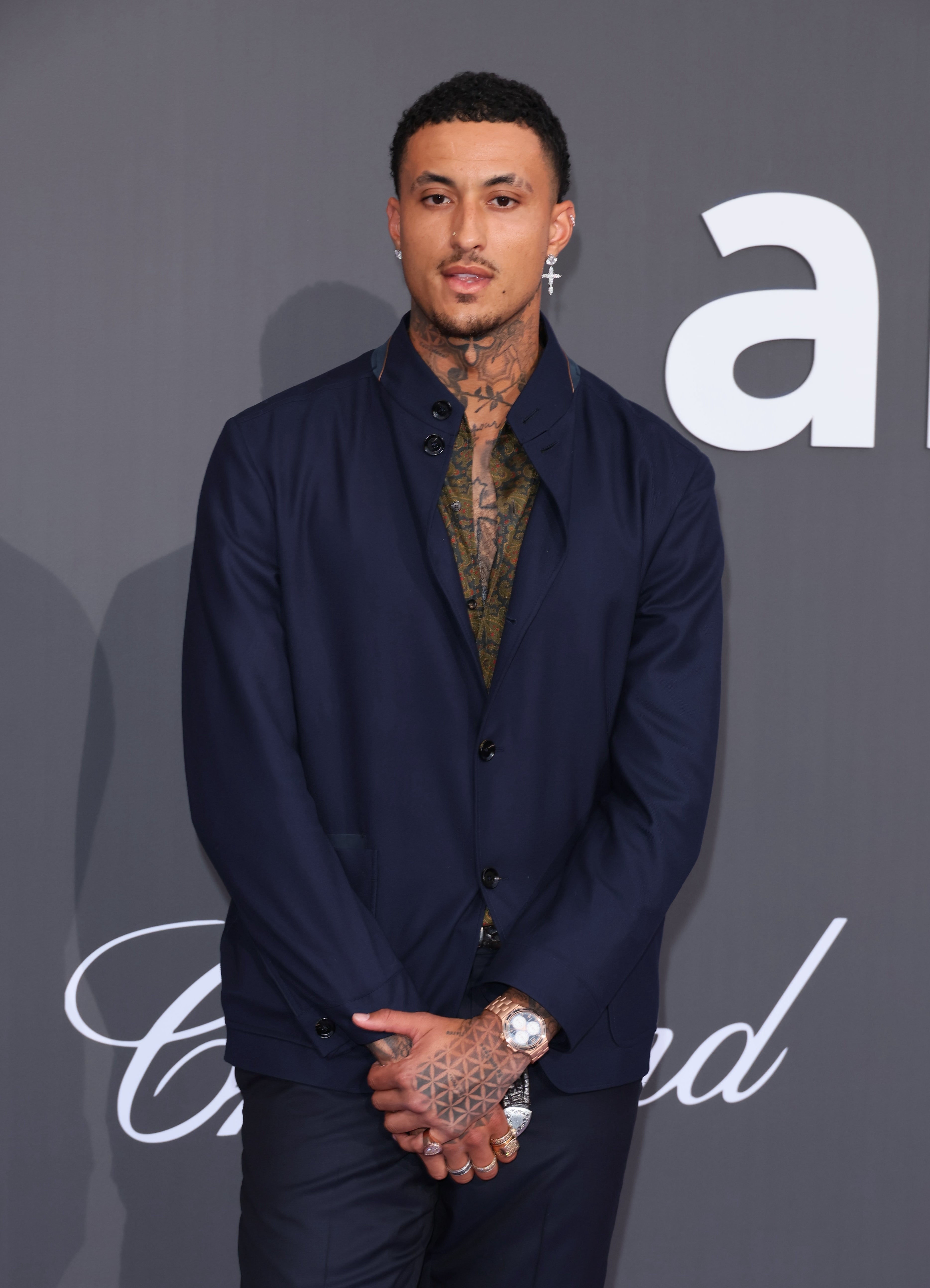 Person in a tailored suit with a patterned shirt underneath, standing at an event. Hands clasped; watch and tattoo visible