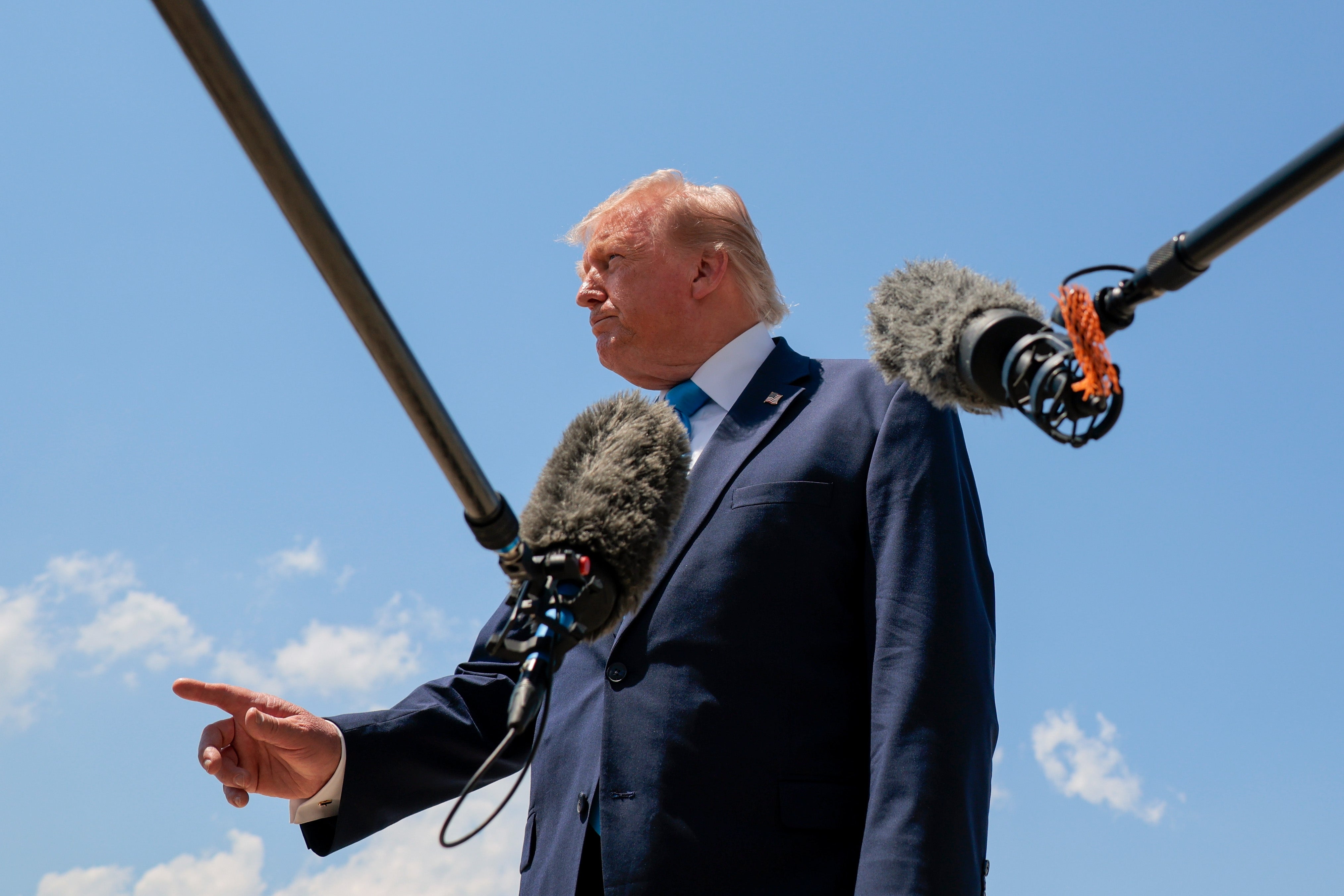 Donald Trump in a suit gestures while surrounded by microphones outdoors, suggesting a press event