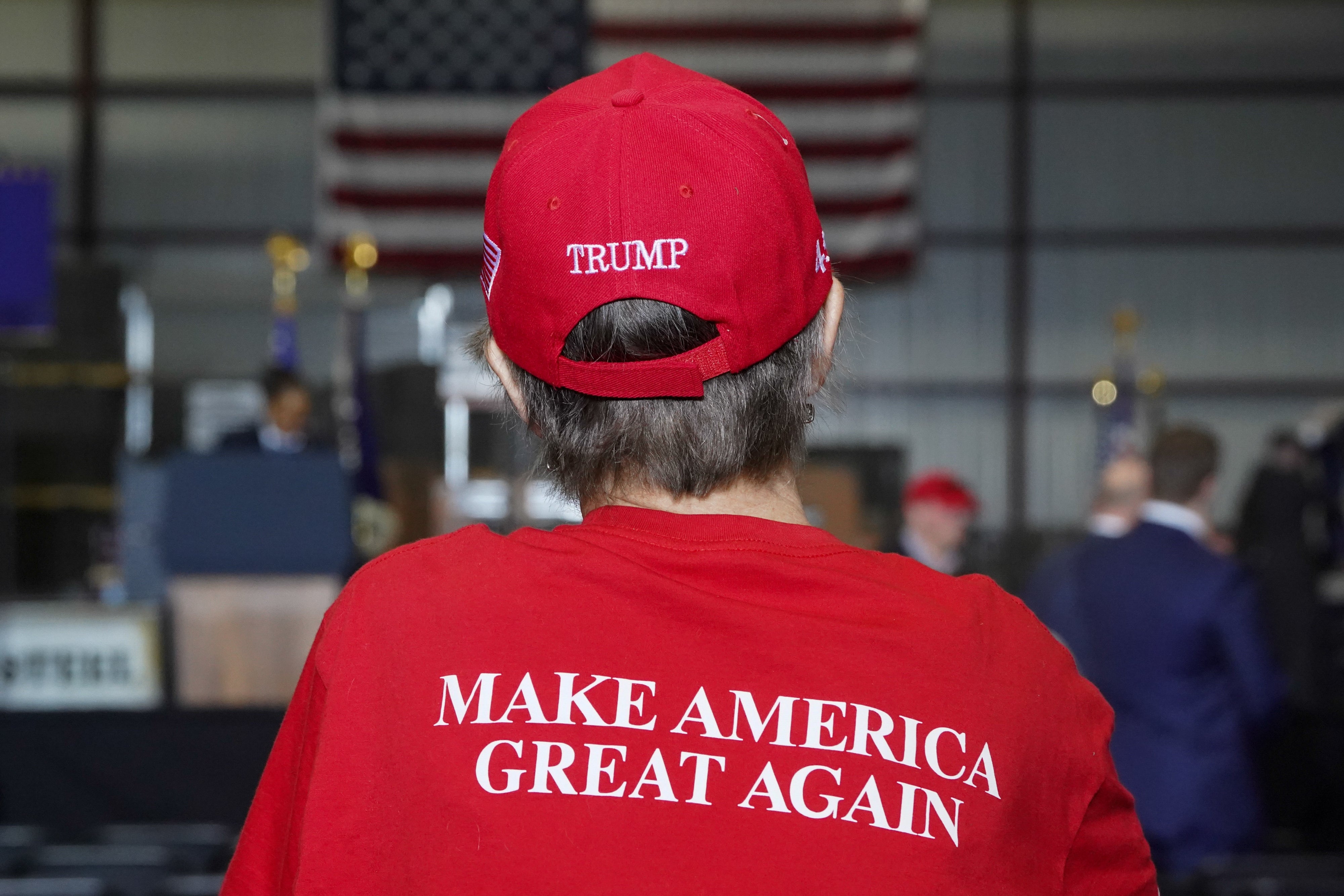 Person wearing a red "Make America Great Again" hat and shirt at an event with U.S. flags in the background