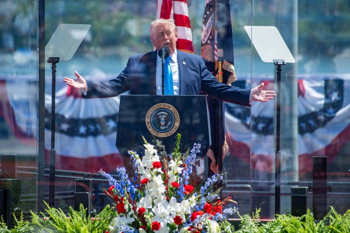 Donald Trump at a podium with U.S. flags and patriotic decorations, gesturing widely, delivering a speech behind glass