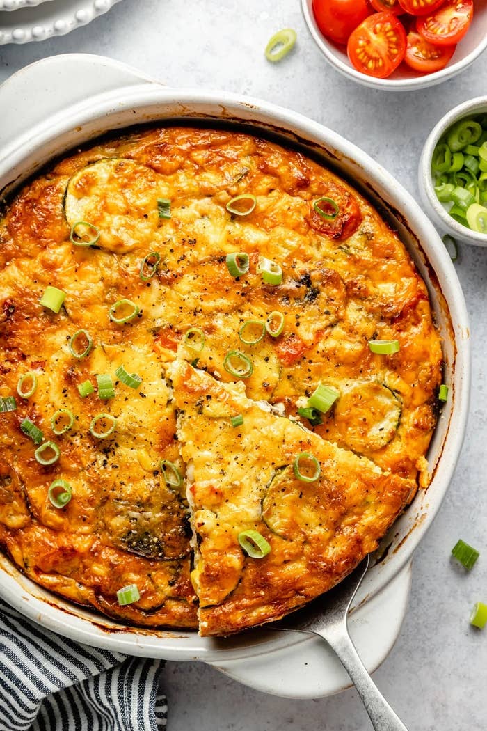 Baked vegetable quiche in a round dish with a slice missing, garnished with scallions. Cherry tomatoes and extra scallions on the side