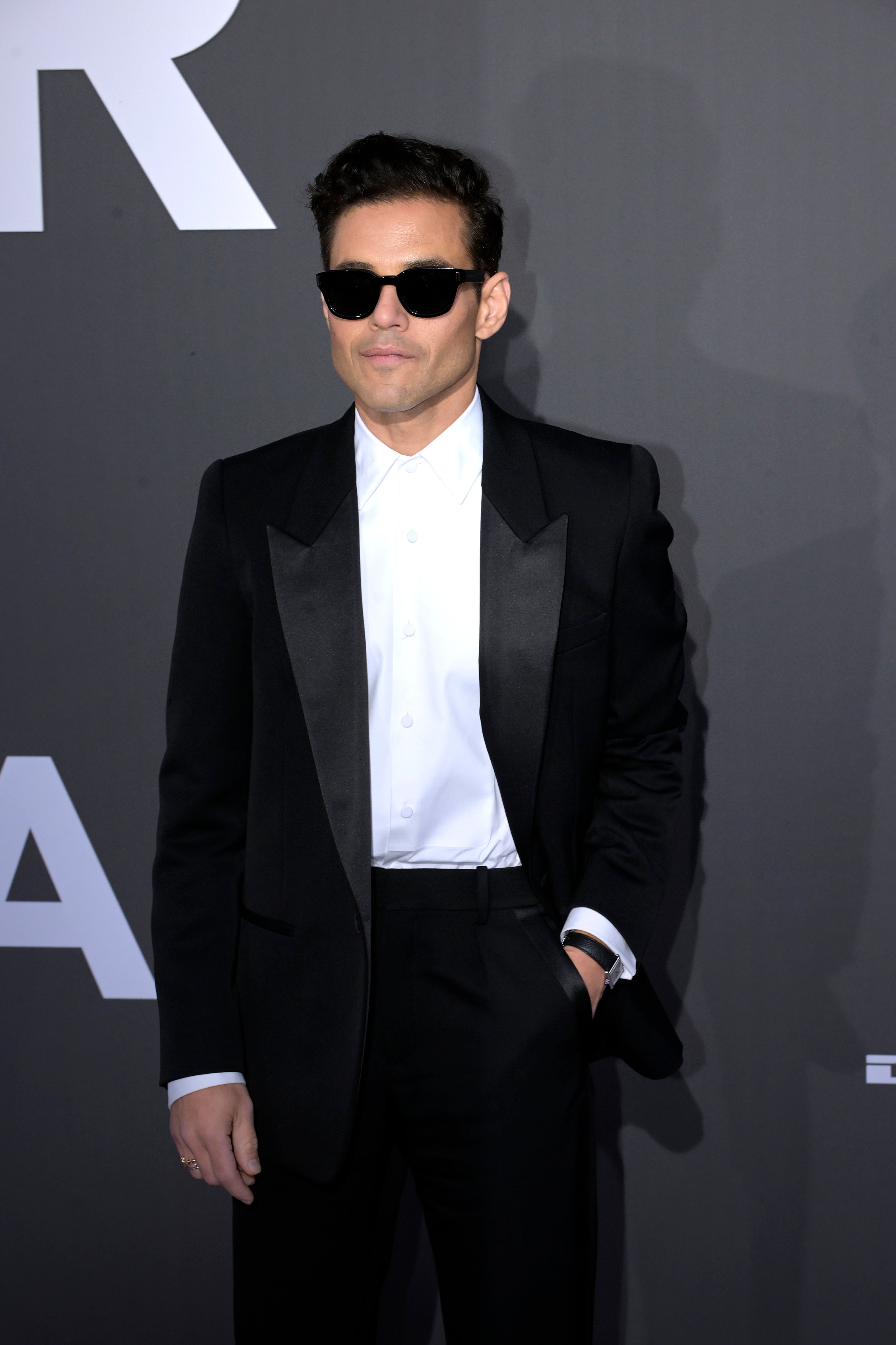 Person in a sleek tuxedo with sunglasses poses confidently on a red carpet