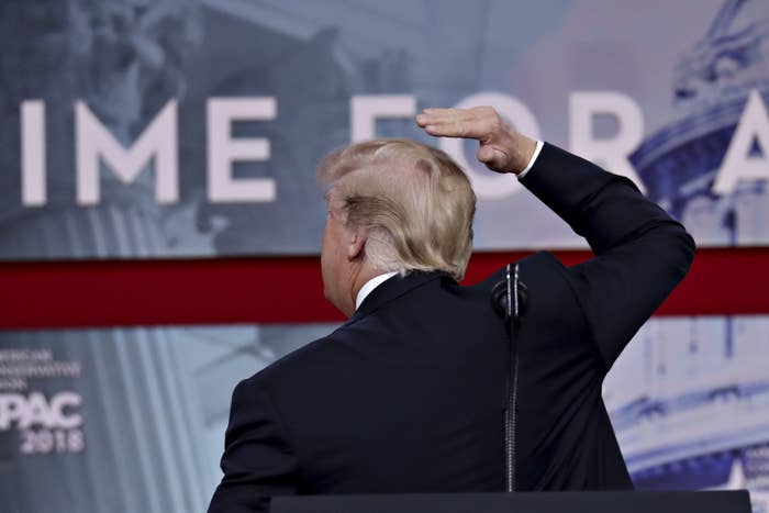 Donald Trump on stage, seen from behind, gesturing to hair. Background includes indistinct signage and architecture. Associated with event presentation
