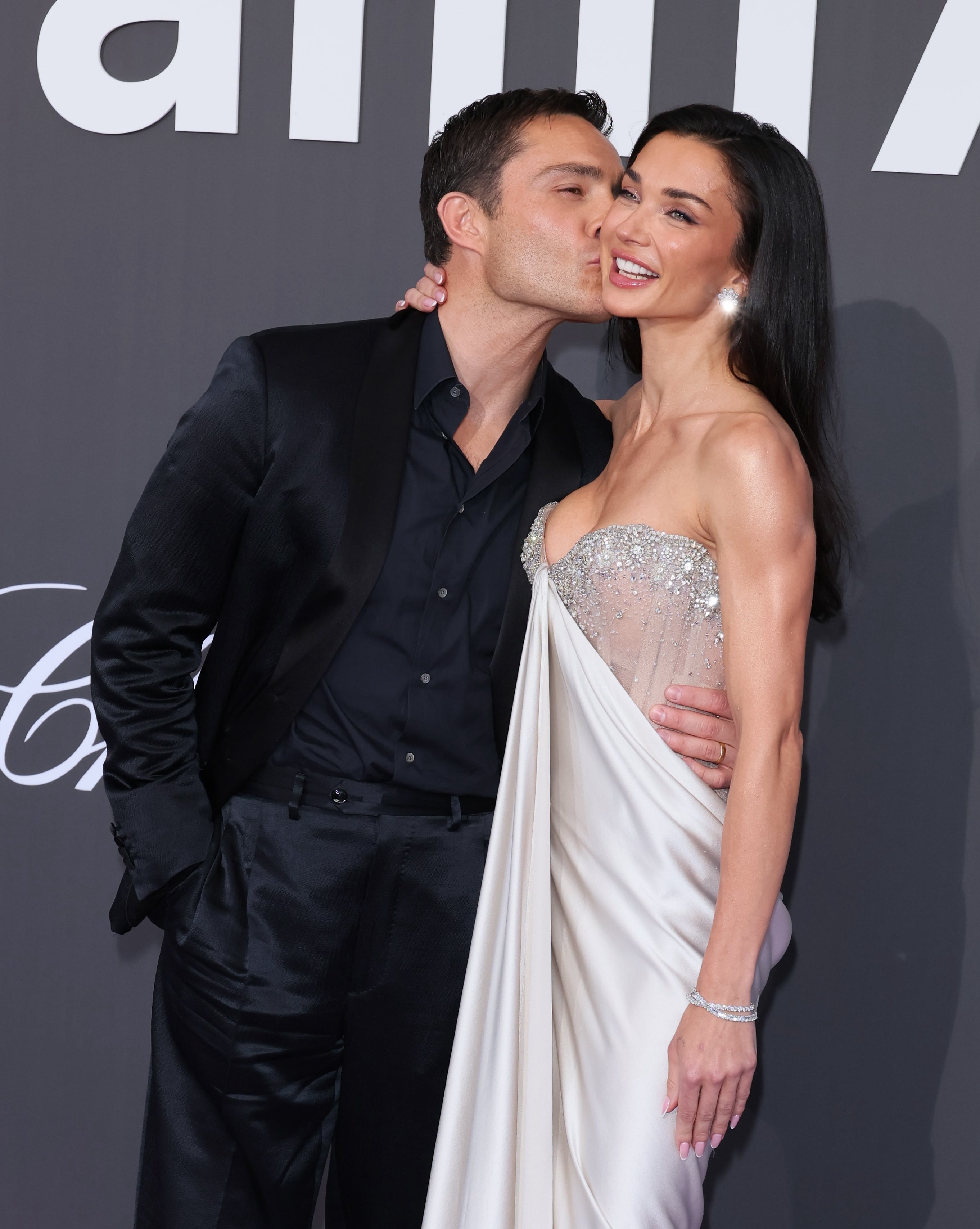 Man in sleek suit kisses woman's cheek; she's in an elegant strapless gown, adorned with jewels, on a red carpet