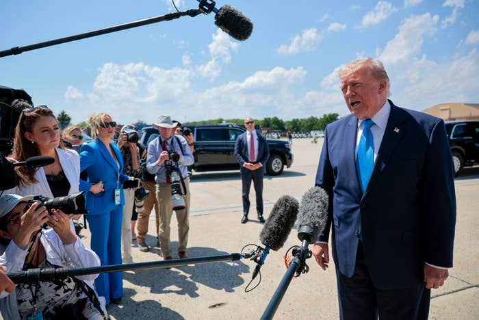 Donald Trump in a suit speaks to the press outdoors, surrounded by microphones and photographers