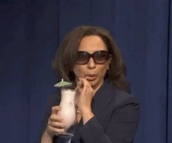 A person with long hair, wearing sunglasses, sipping a drink with an umbrella while making a playful expression