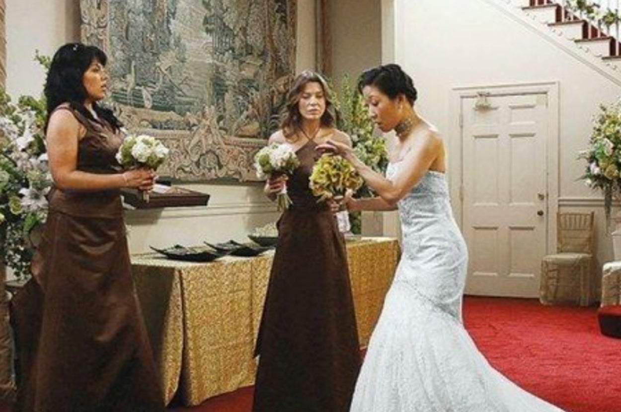Three women in a wedding scene. Two in matching gowns hold bouquets, while the one in a bridal gown interacts with them. Elegant setting