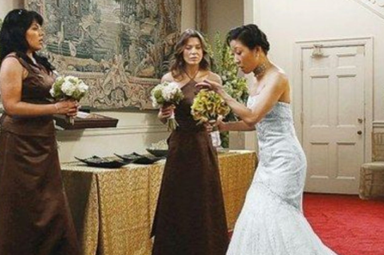 Three women in a wedding scene. Two in matching gowns hold bouquets, while the one in a bridal gown interacts with them. Elegant setting