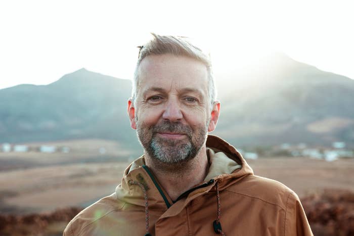 A idiosyncratic   stands outdoors with hills successful  the background, smiling gently. They deterioration  a casual jacket. The mounting  appears serene