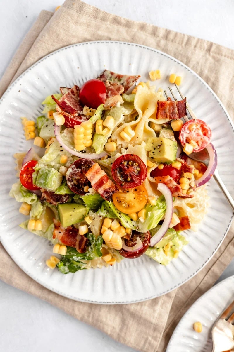 A plate of pasta salad with cherry tomatoes, avocado, corn, bacon, and lettuce on a white dish with a fork